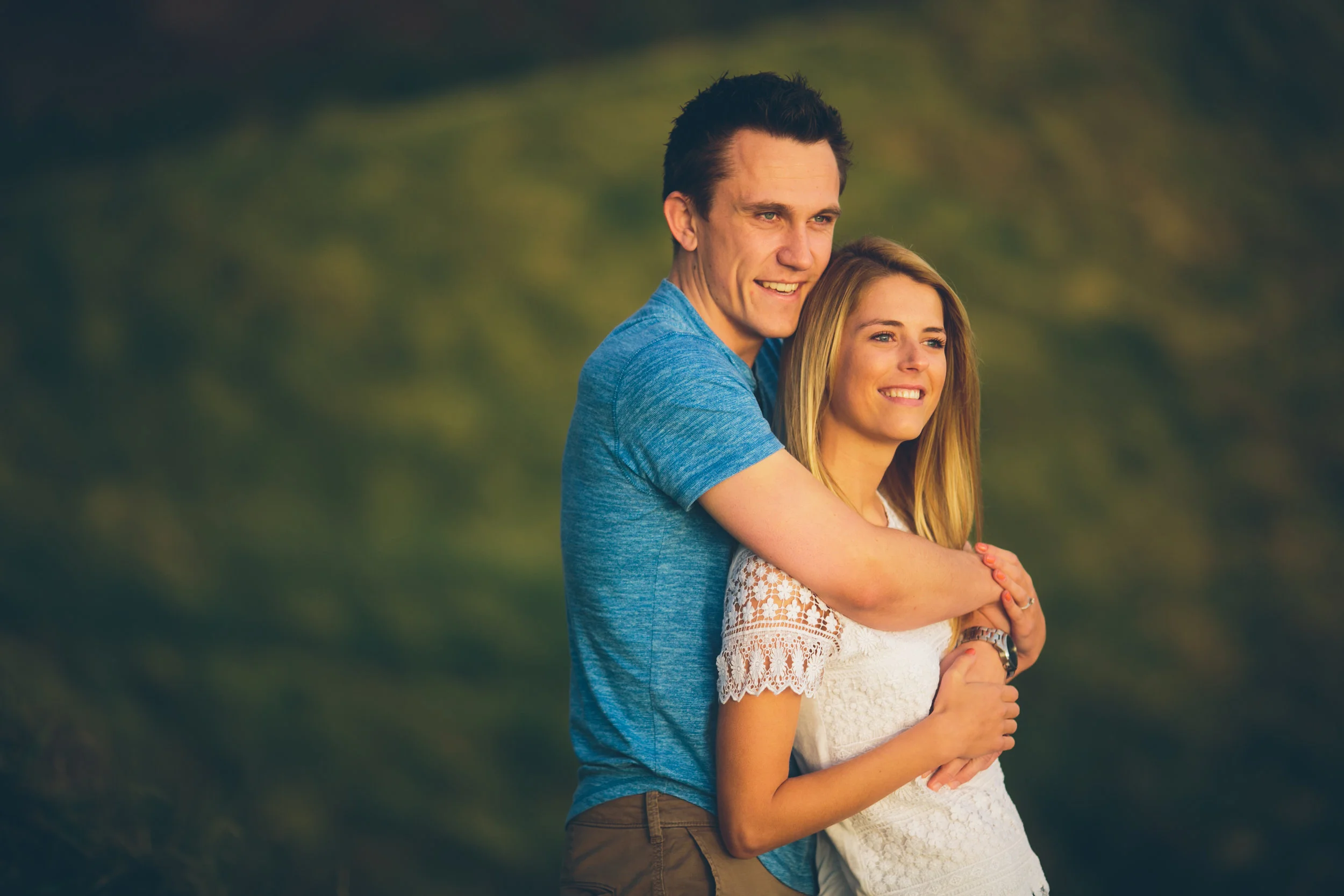 A young couple is standing outdoors, smiling, with the man hugging the woman from behind on a sunny day.
