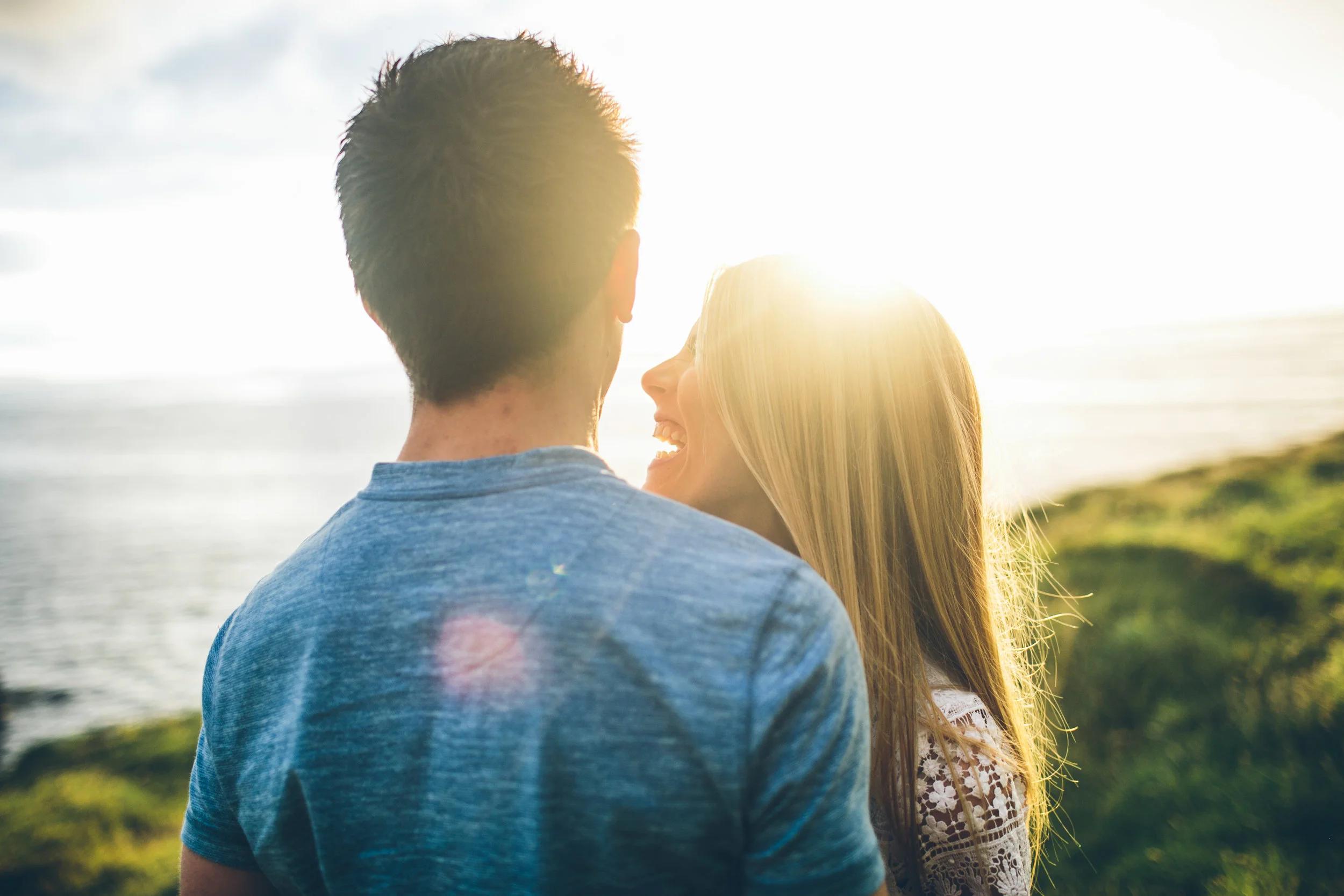 A young couple standing close together outdoors near the ocean during sunset, smiling and enjoying each other's company.