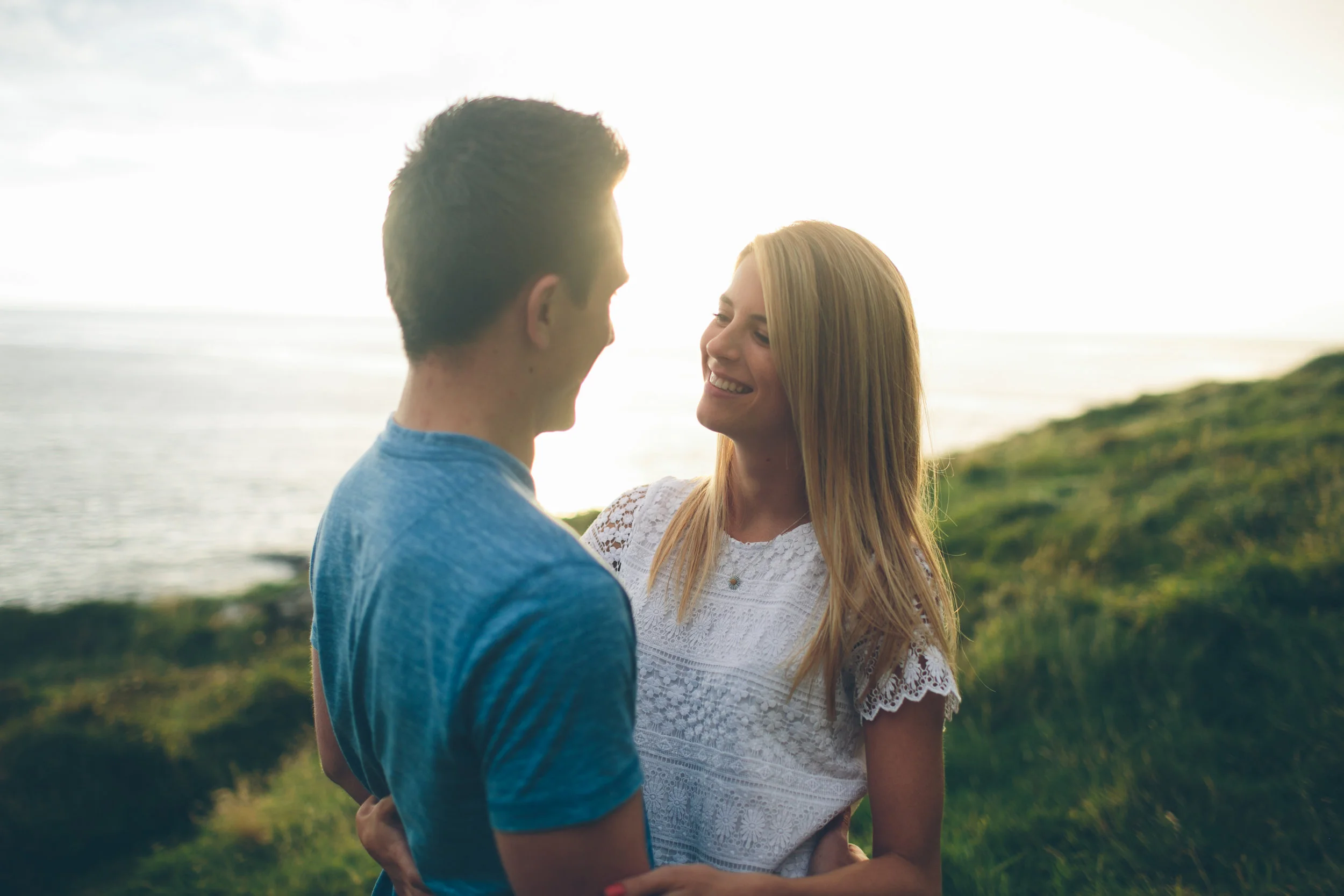 A young couple standing close together outdoors near the water, smiling and looking at each other during sunset.