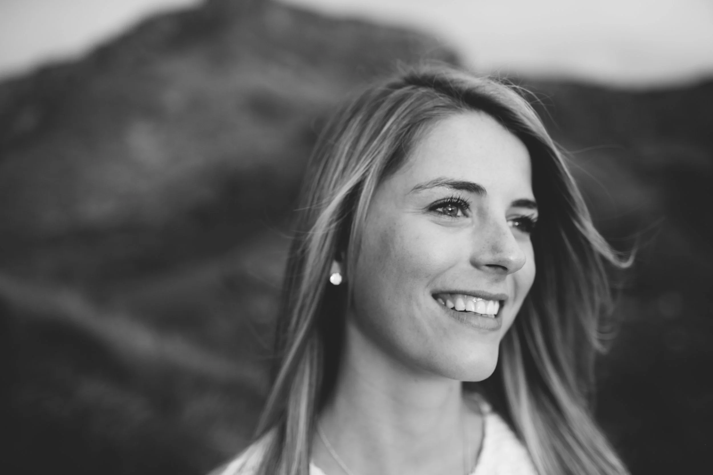 A smiling woman with long hair outdoors with mountains in the background, in black and white.