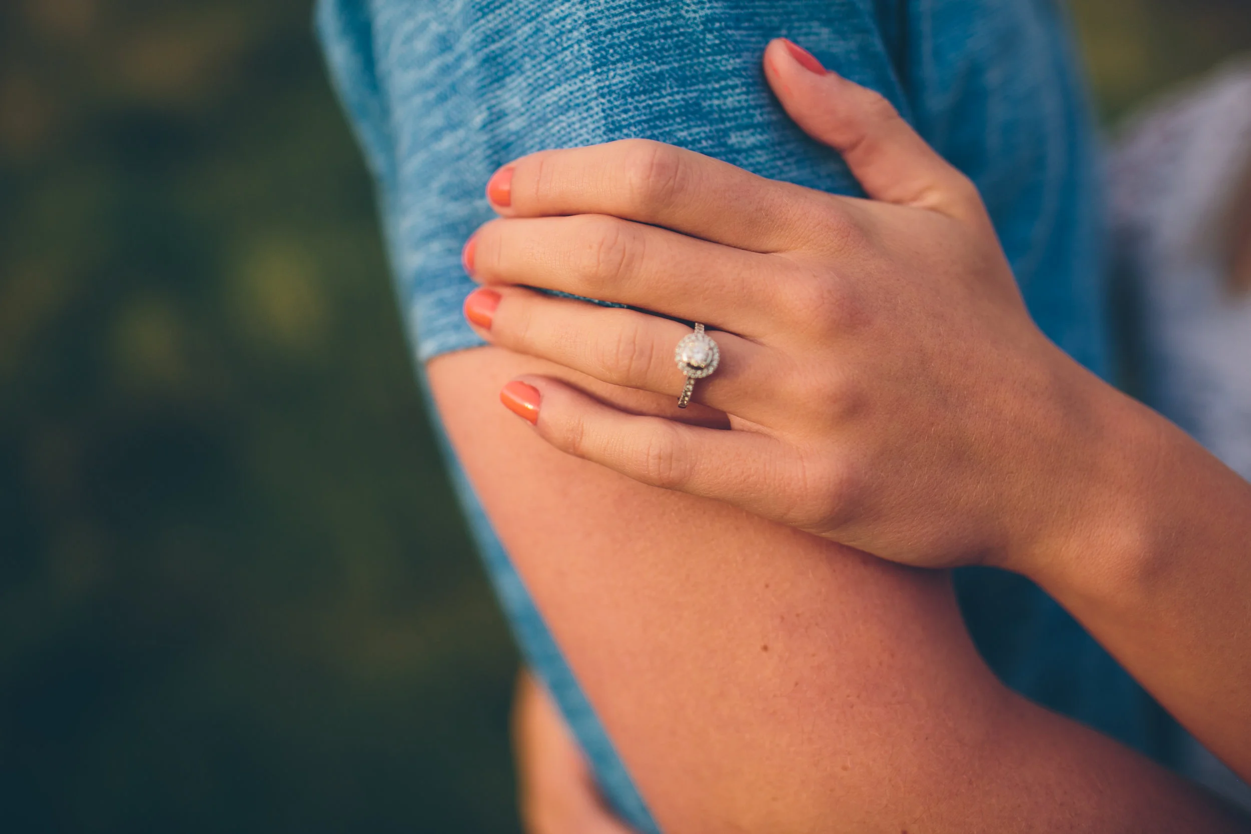 Close-up of a woman's hand displaying a diamond engagement ring on her ring finger, with her other hand resting on her exposed shoulder. She is wearing a blue shirt and has bright coral nail polish.