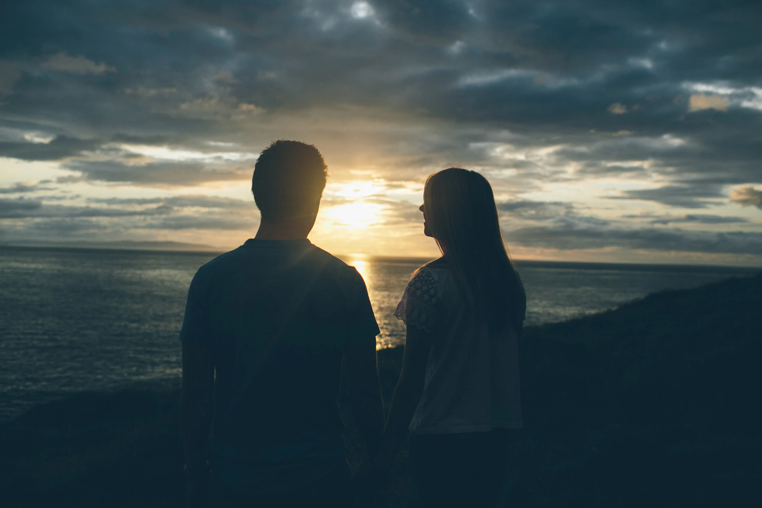A silhouette of a young man and woman holding hands, standing near the coast watching the sunset over the ocean with cloudy skies.