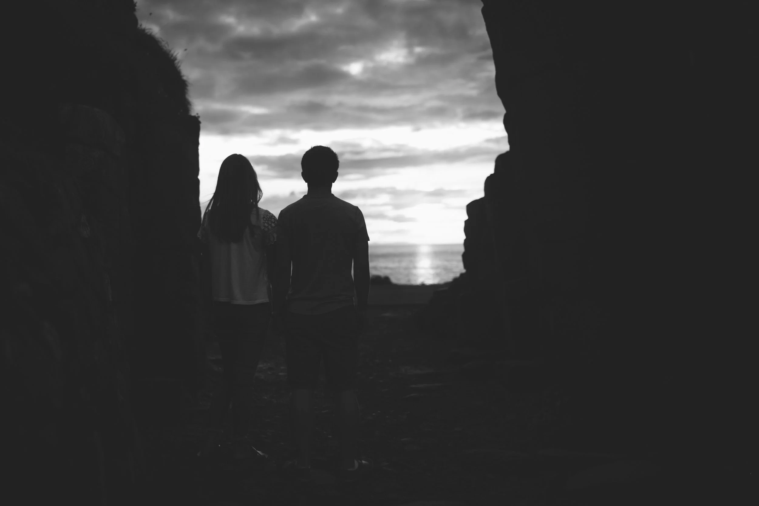 A silhouette of a girl and a boy walking hand in hand through a rocky opening, towards the sunset view of the ocean and cloudy sky.