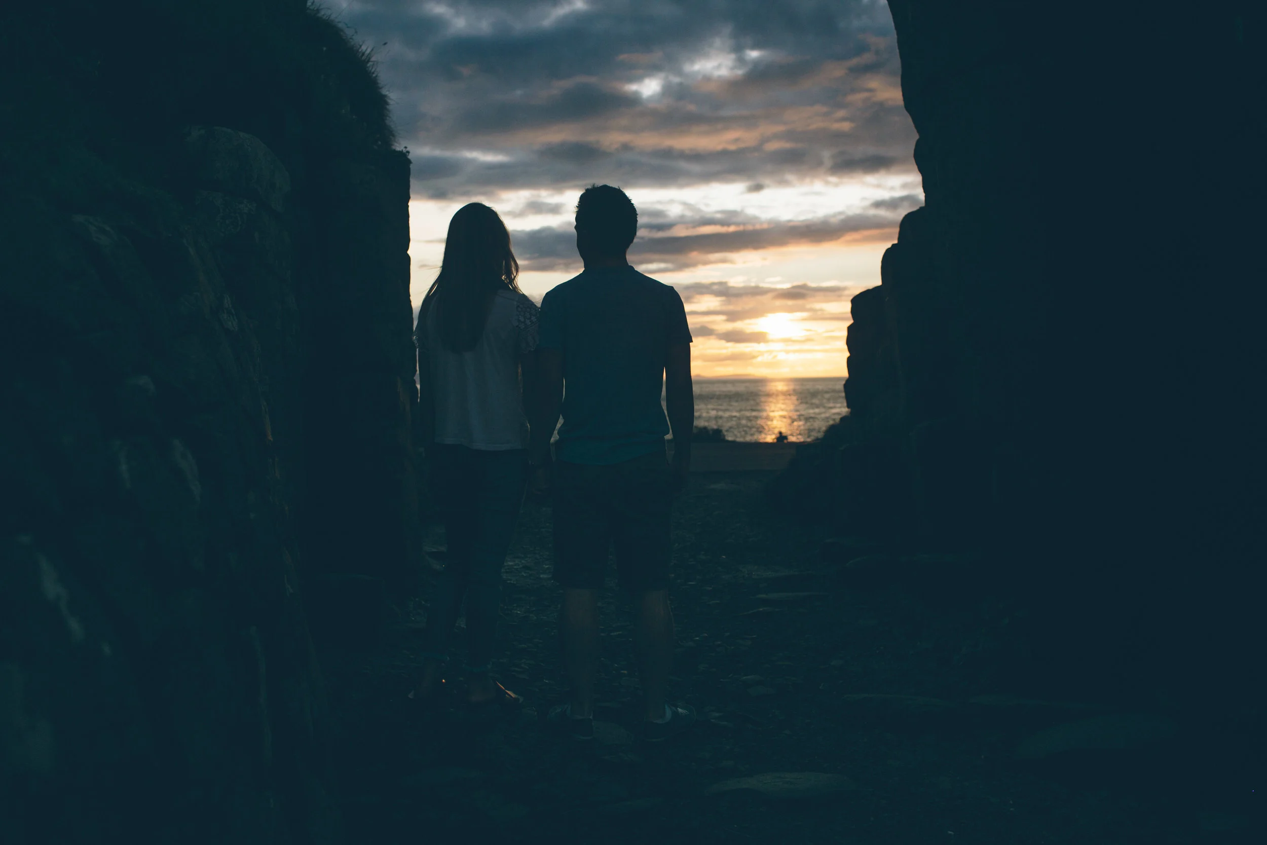 A silhouette of a couple holding hands, standing inside a rocky cave opening, overlooking the ocean at sunset with cloudy skies.