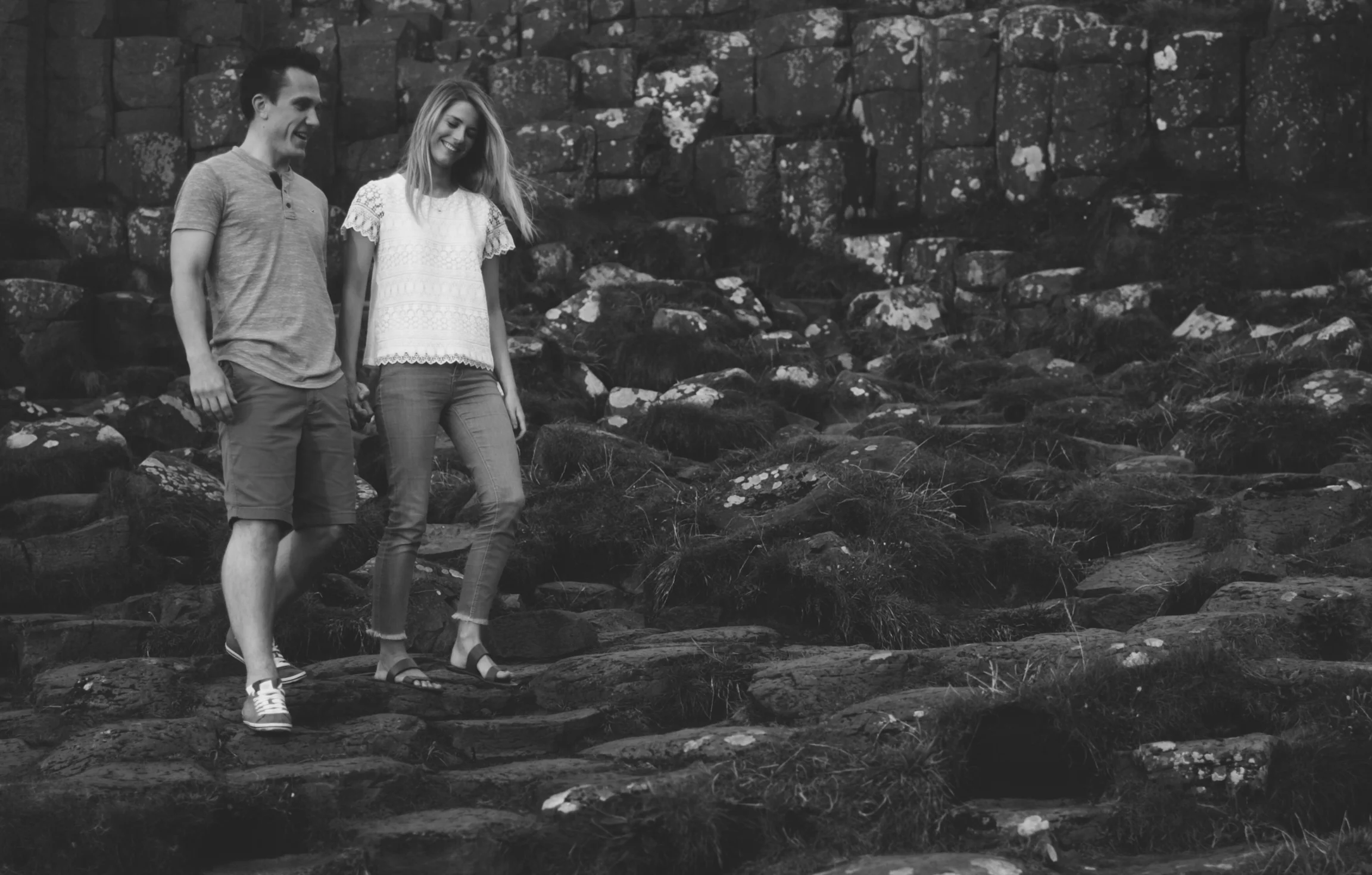 A black-and-white photo of a young man and woman walking on rocks outdoors, with a stone wall in the background. They are holding hands and smiling.