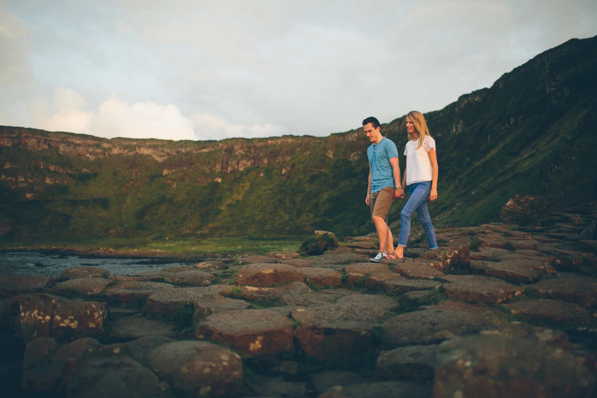A young man and woman walking on a rocky path near the water in a green mountainous landscape during daylight.