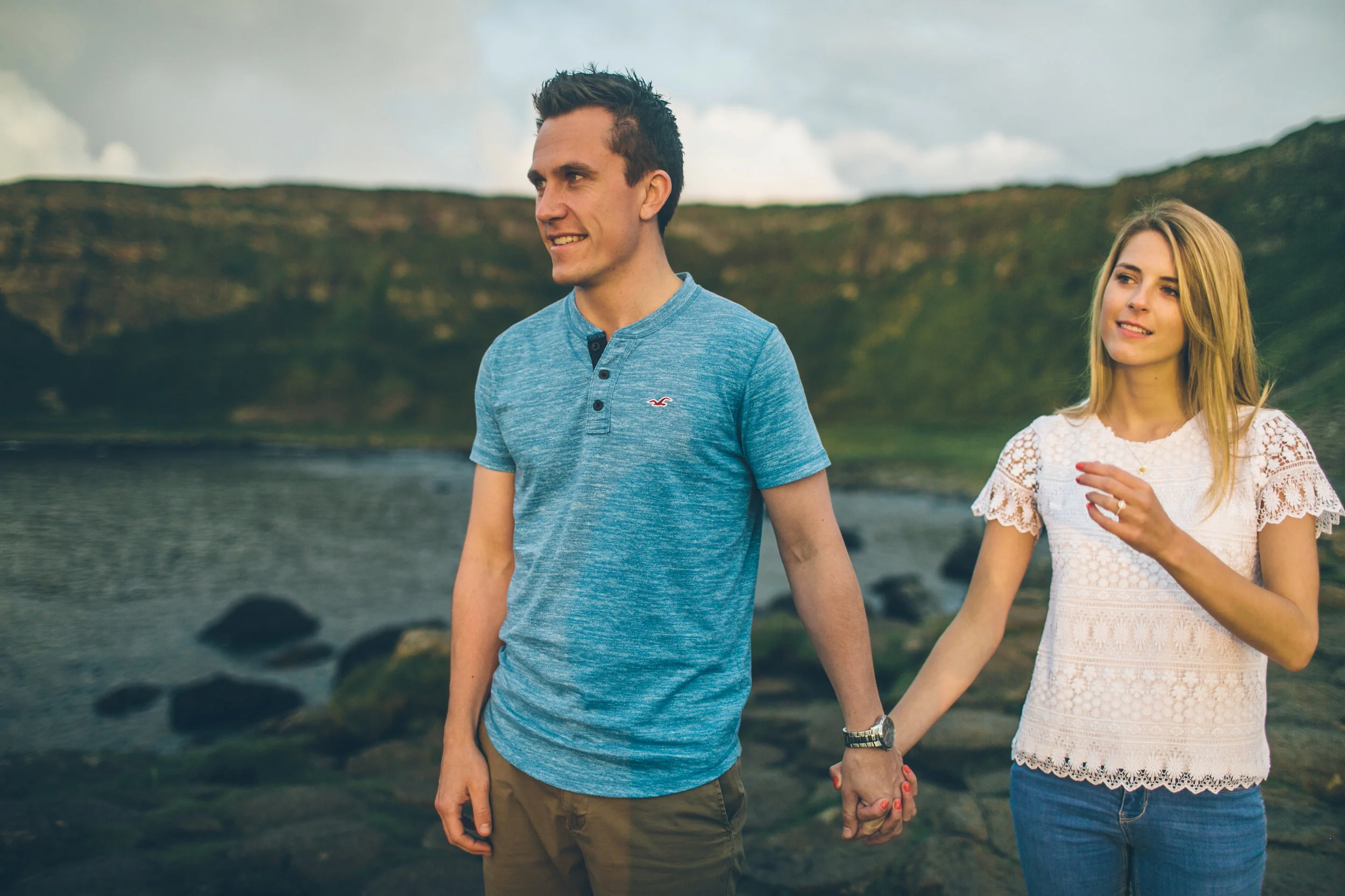 A man and a woman holding hands on a rocky shoreline with mountains and water in the background.