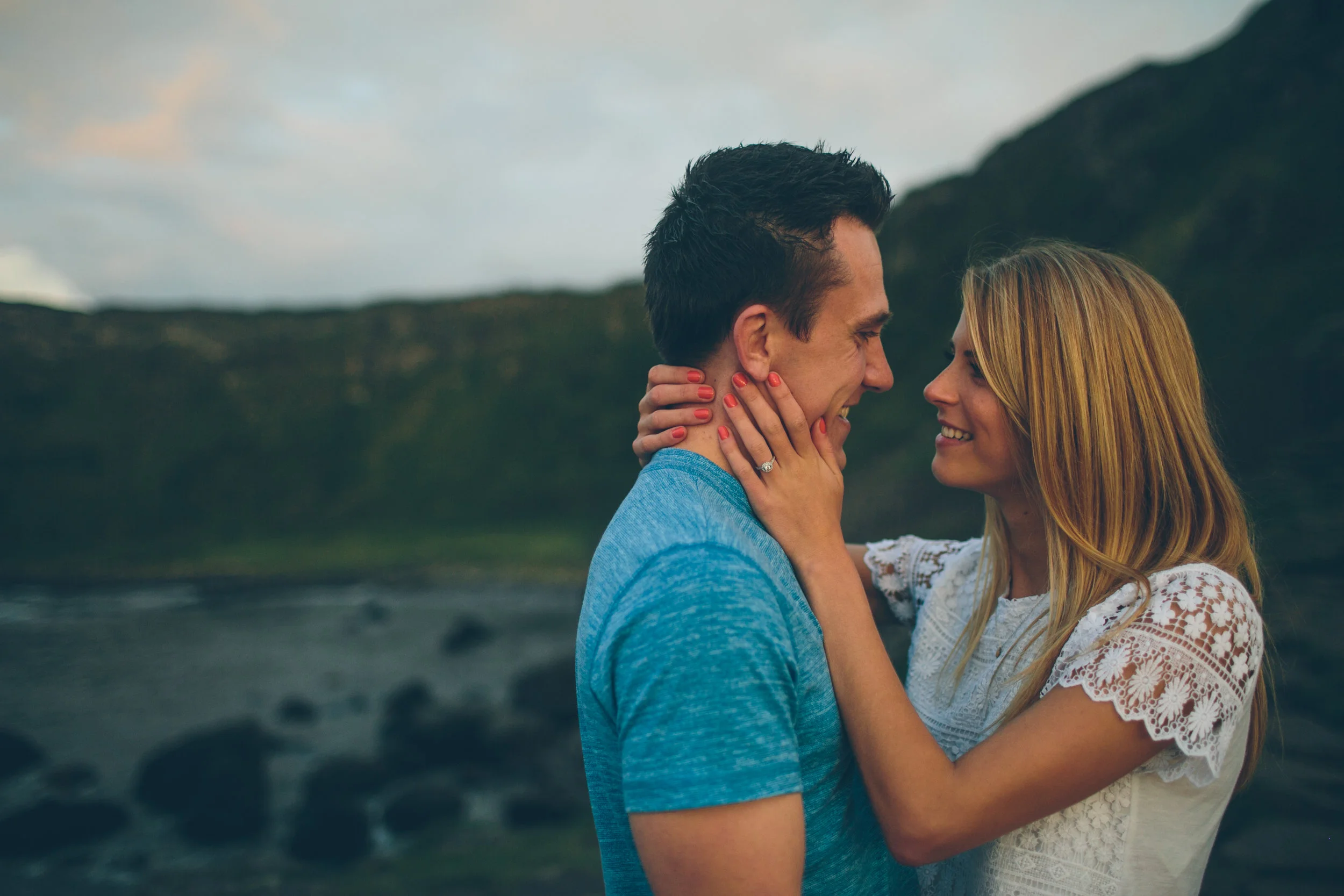 A man and woman smiling and looking at each other outdoors, with hills and a cloudy sky in the background.