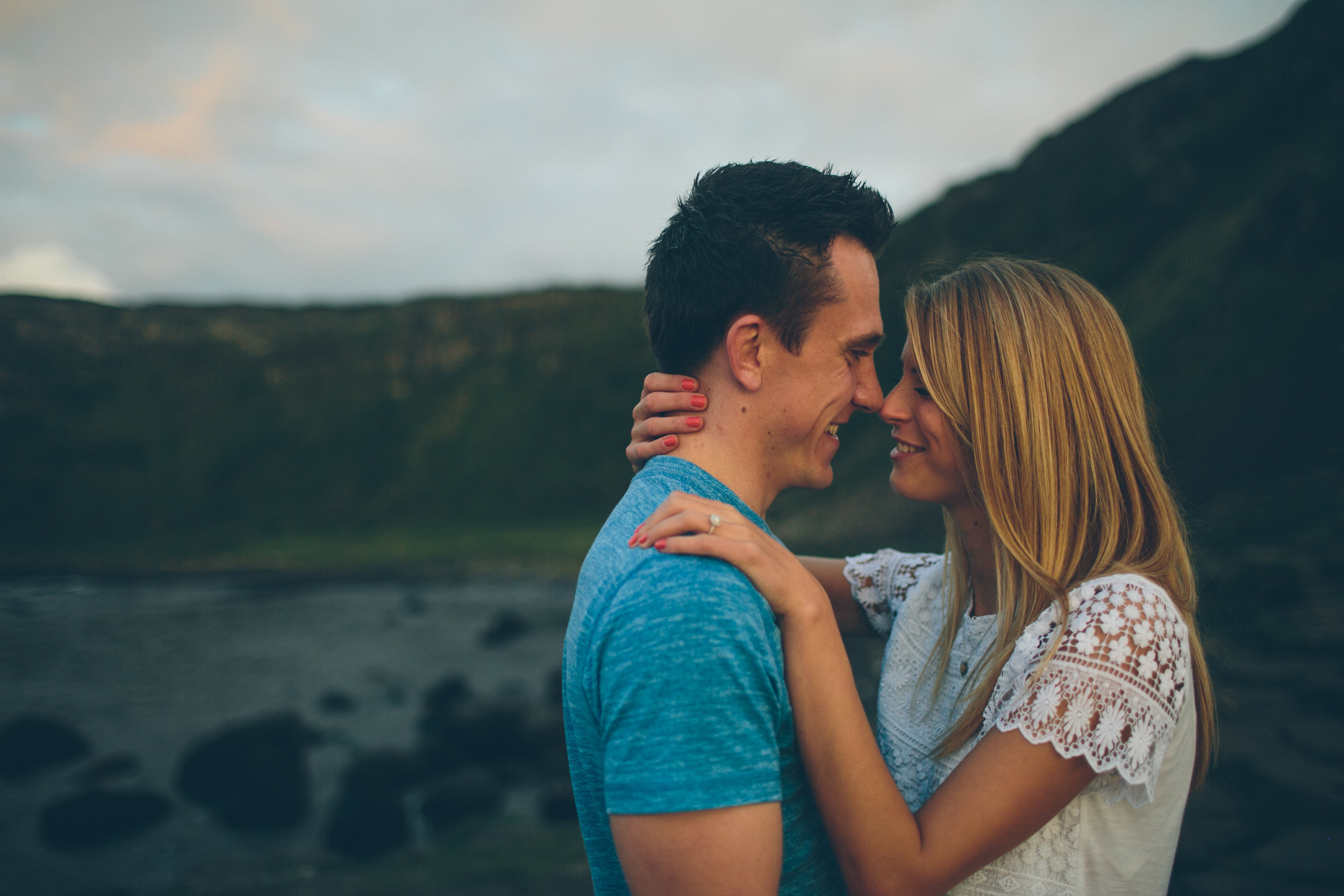 A couple embracing outdoors, smiling at each other with a mountain and cloudy sky in the background.