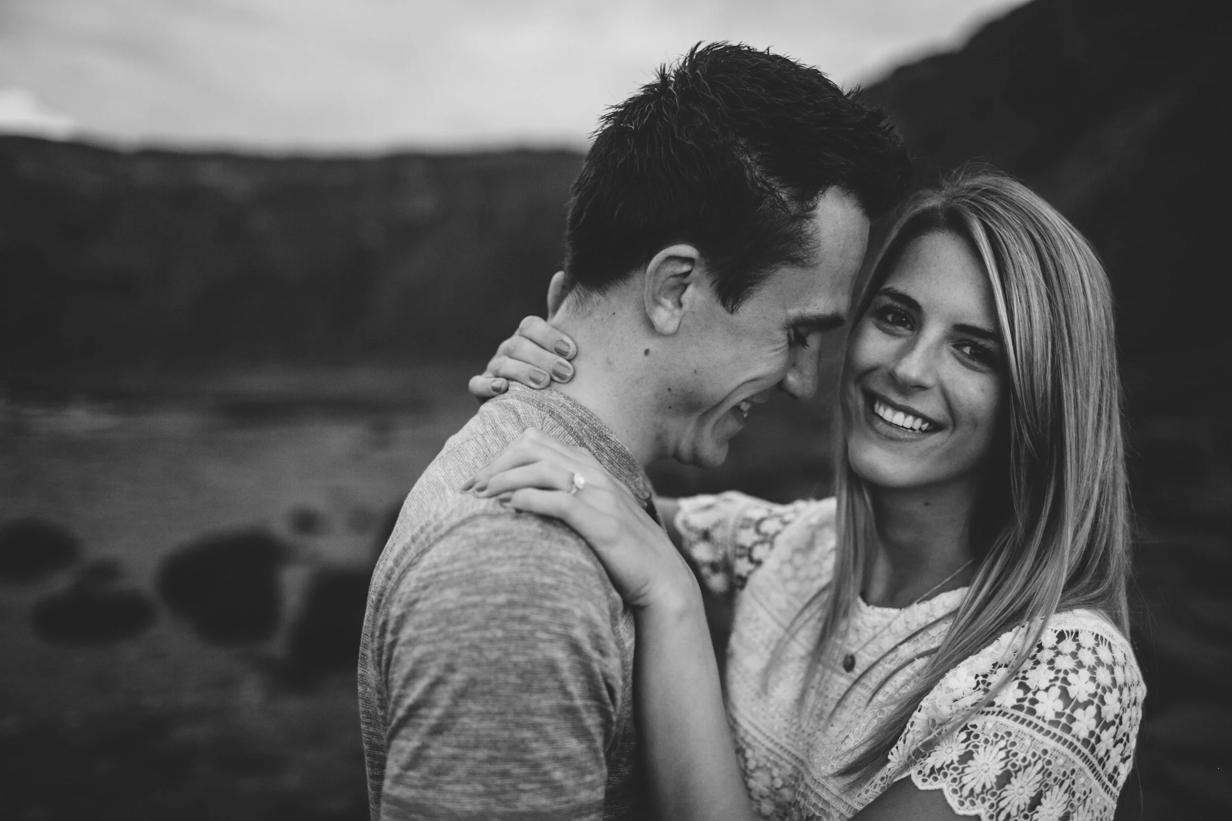 A black-and-white photo of a smiling couple standing close together outdoors, with a mountainous landscape in the background. The woman has long hair and is wearing a lace dress, and the man has short hair and is wearing a striped shirt.