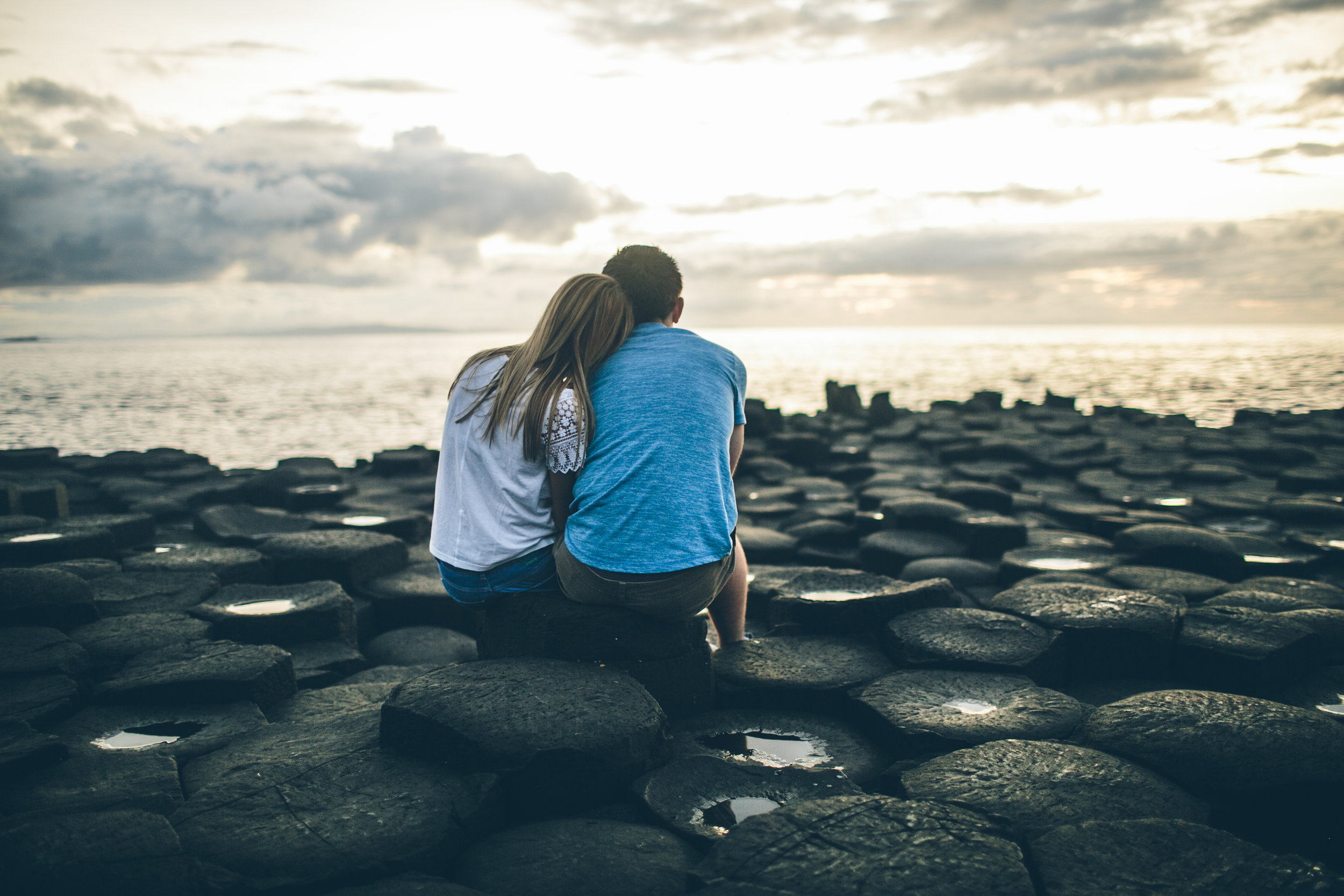 A couple sitting on rocks by the water at sunset, with the ocean and cloudy sky in the background.