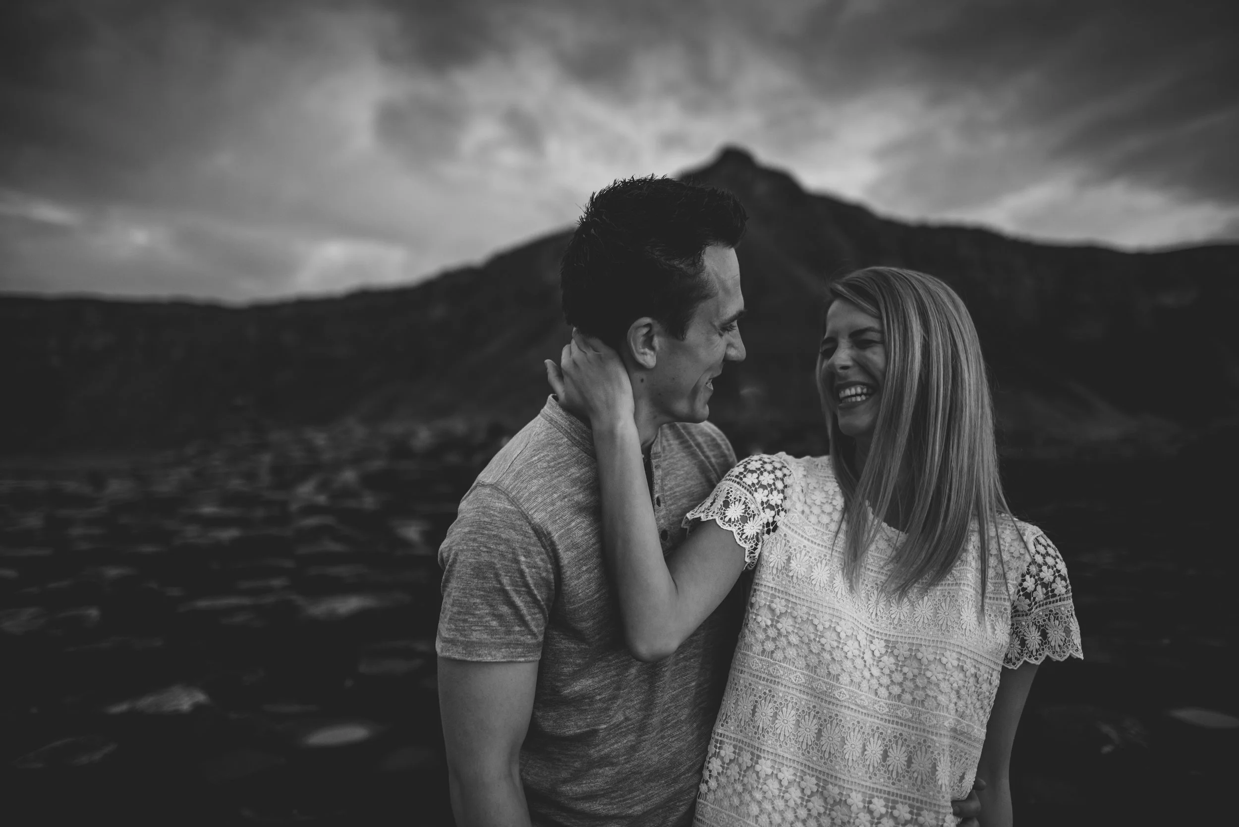 A couple smiling and looking at each other, standing outdoors with a mountain and cloudy sky in the background.