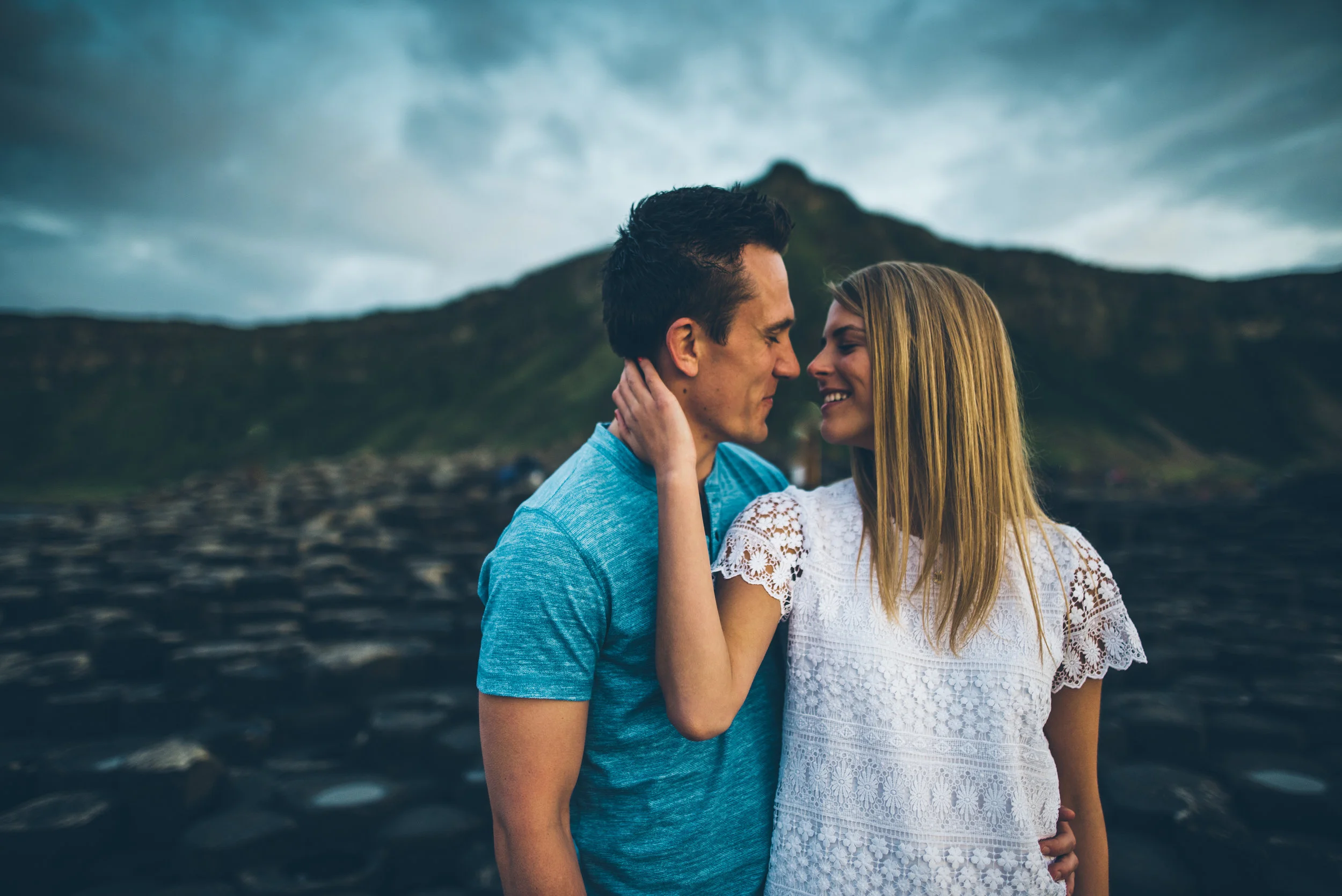 A couple standing close together outdoors with lush green hills and cloudy sky in the background.