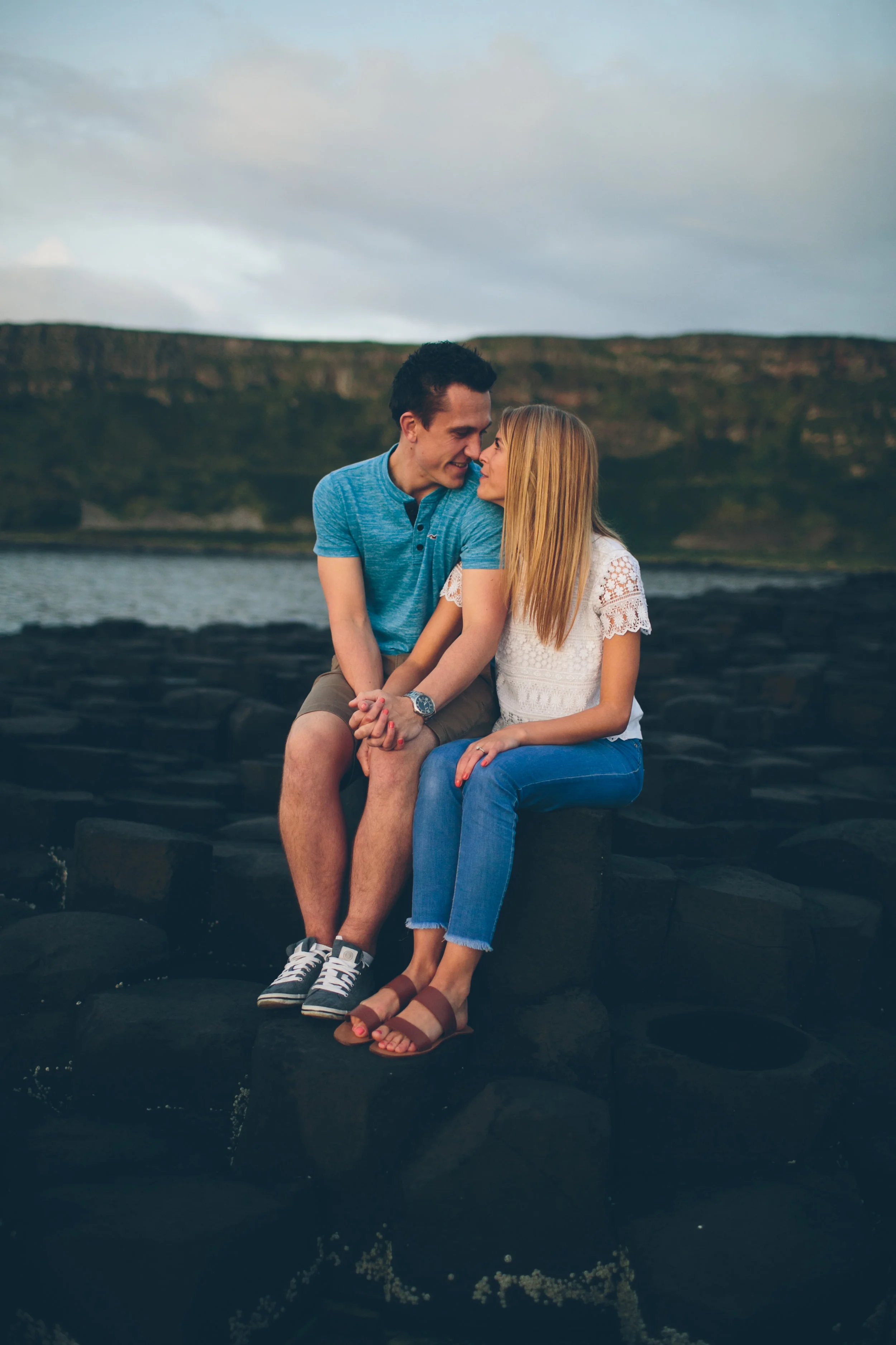 A young couple sitting close together on dark rocks near a body of water, sharing a tender moment with faces close, outdoors with a cloudy sky and distant hills.