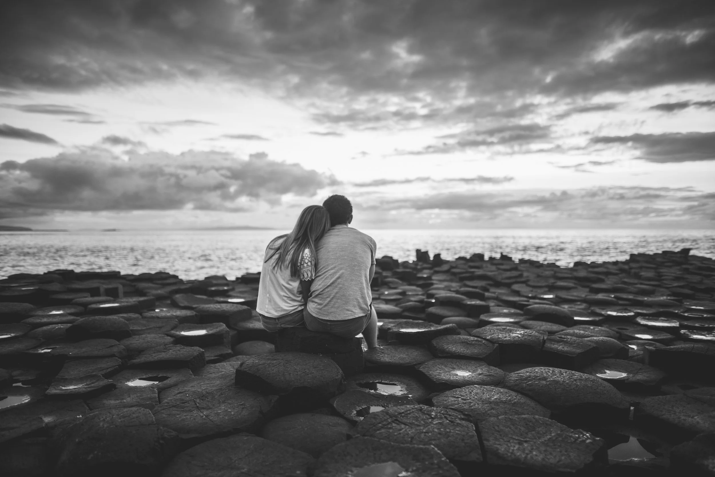 A couple sitting on rocks at the beach, gazing at the ocean during sunset or sunrise in black and white.