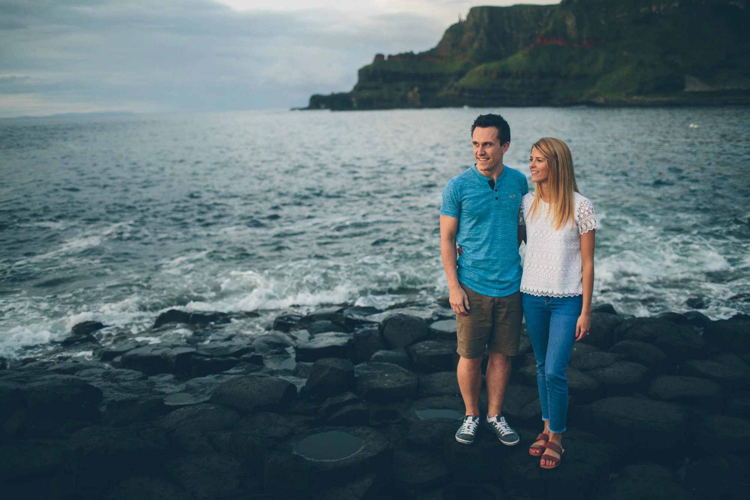 A young man and woman standing on rocks near the ocean, smiling, with a distant green cliff and cloudy sky in the background.