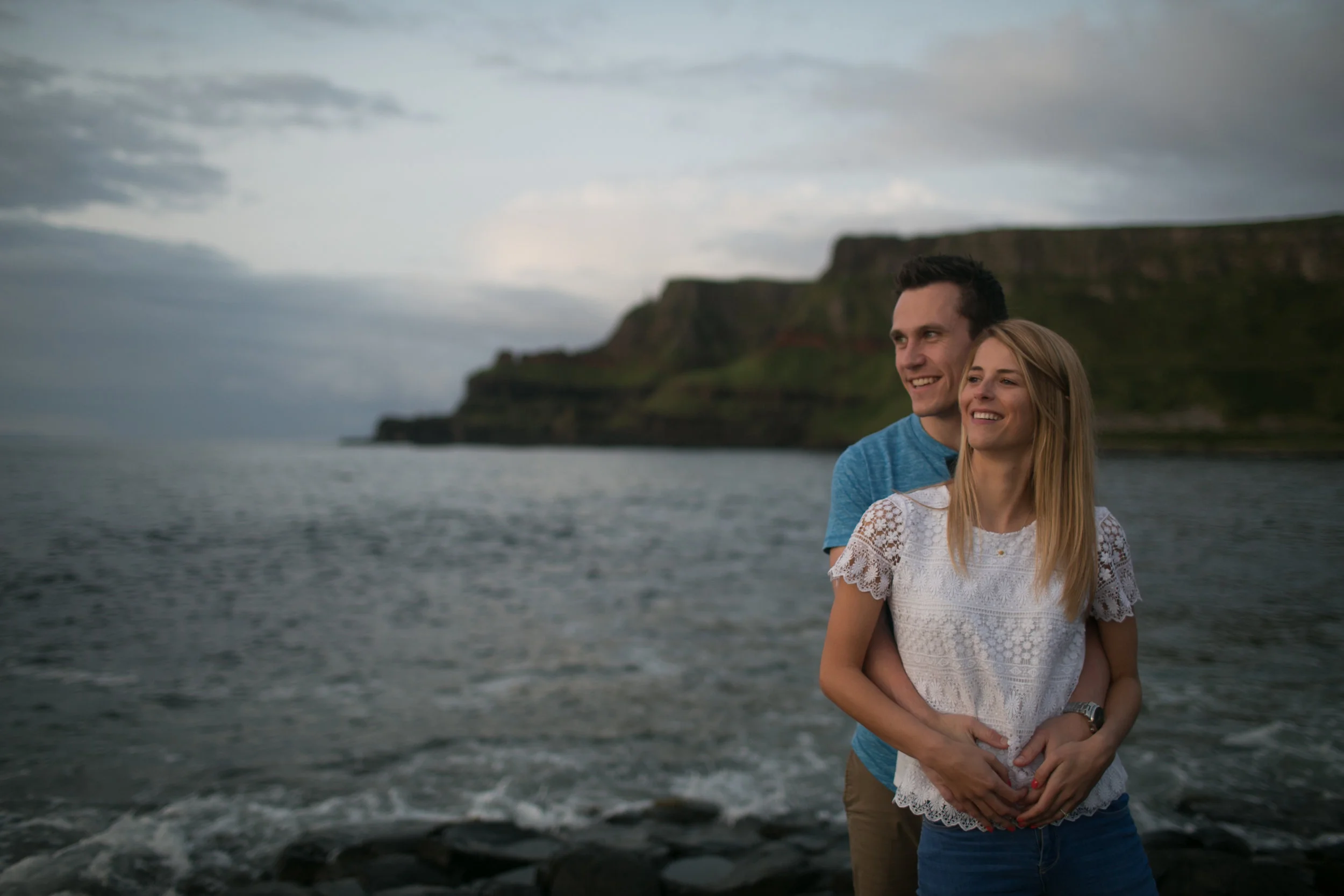 A smiling couple standing on a rocky beach by the water, with green cliffs in the background during an overcast day.