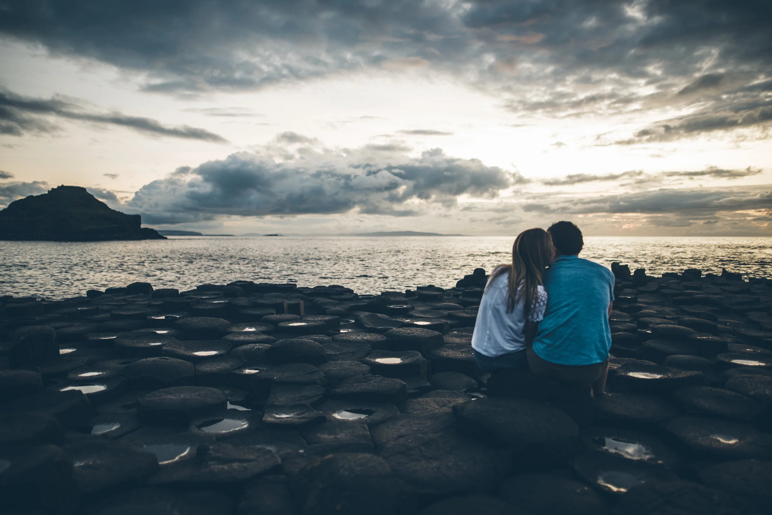 A couple sitting on rocks by the ocean during sunset or sunrise, with cloudy sky and distant landmass.