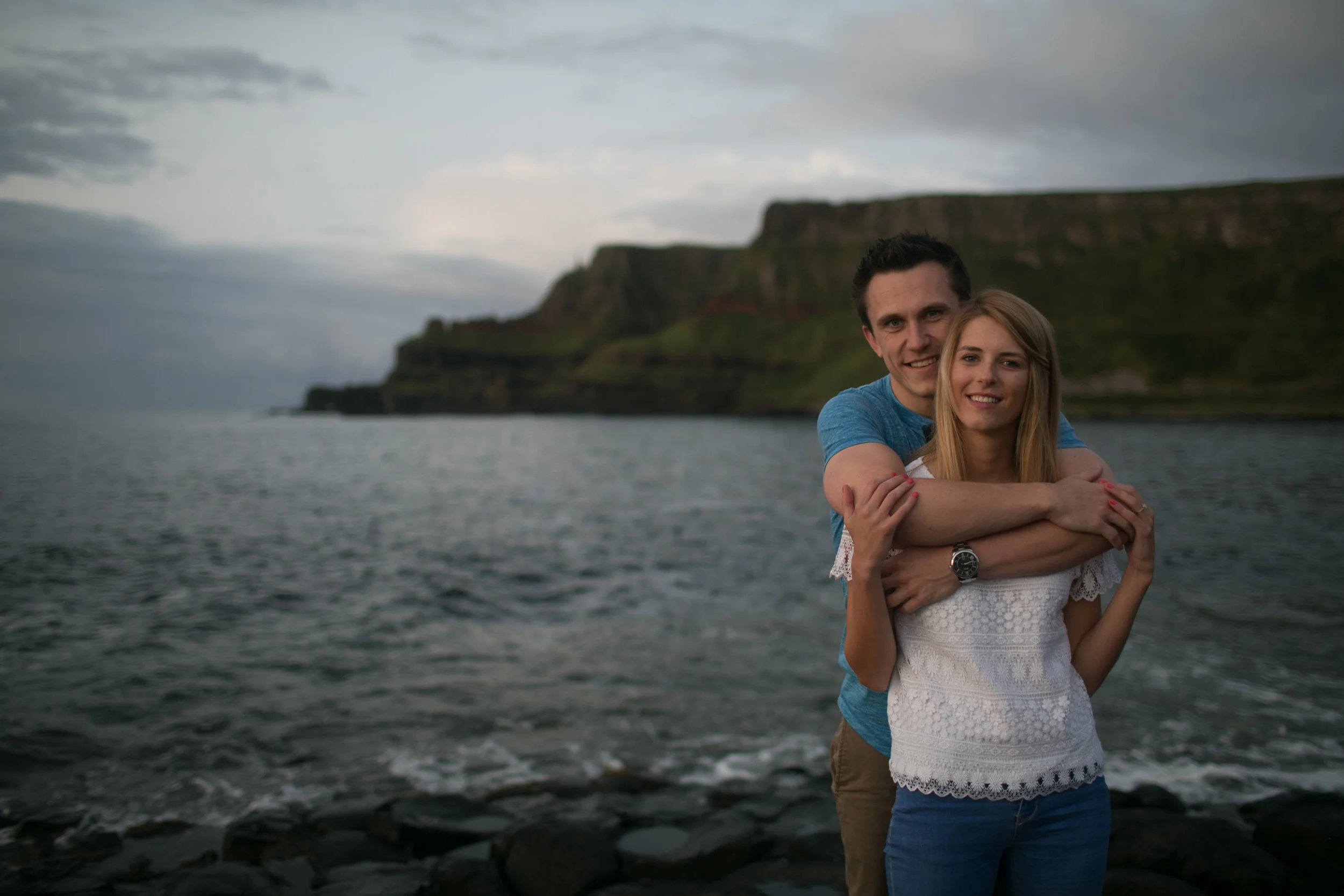 A smiling young couple standing by the water's edge with cliffs in the background, embracing each other.