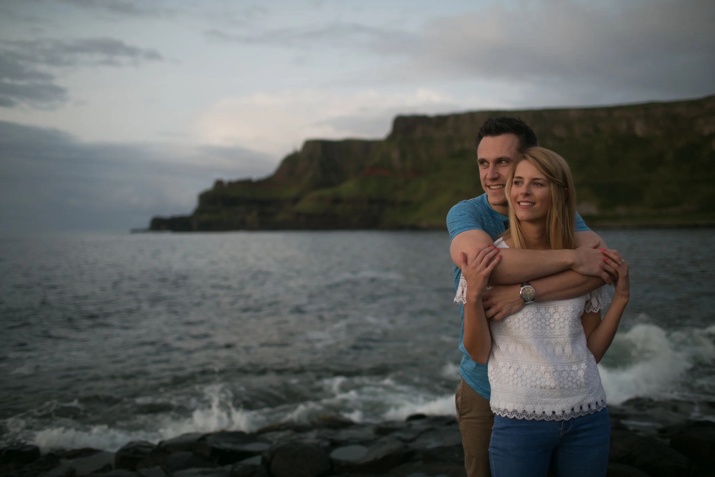 A smiling couple standing on a rocky shoreline embracing with water and cliffs in the background under a cloudy sky.