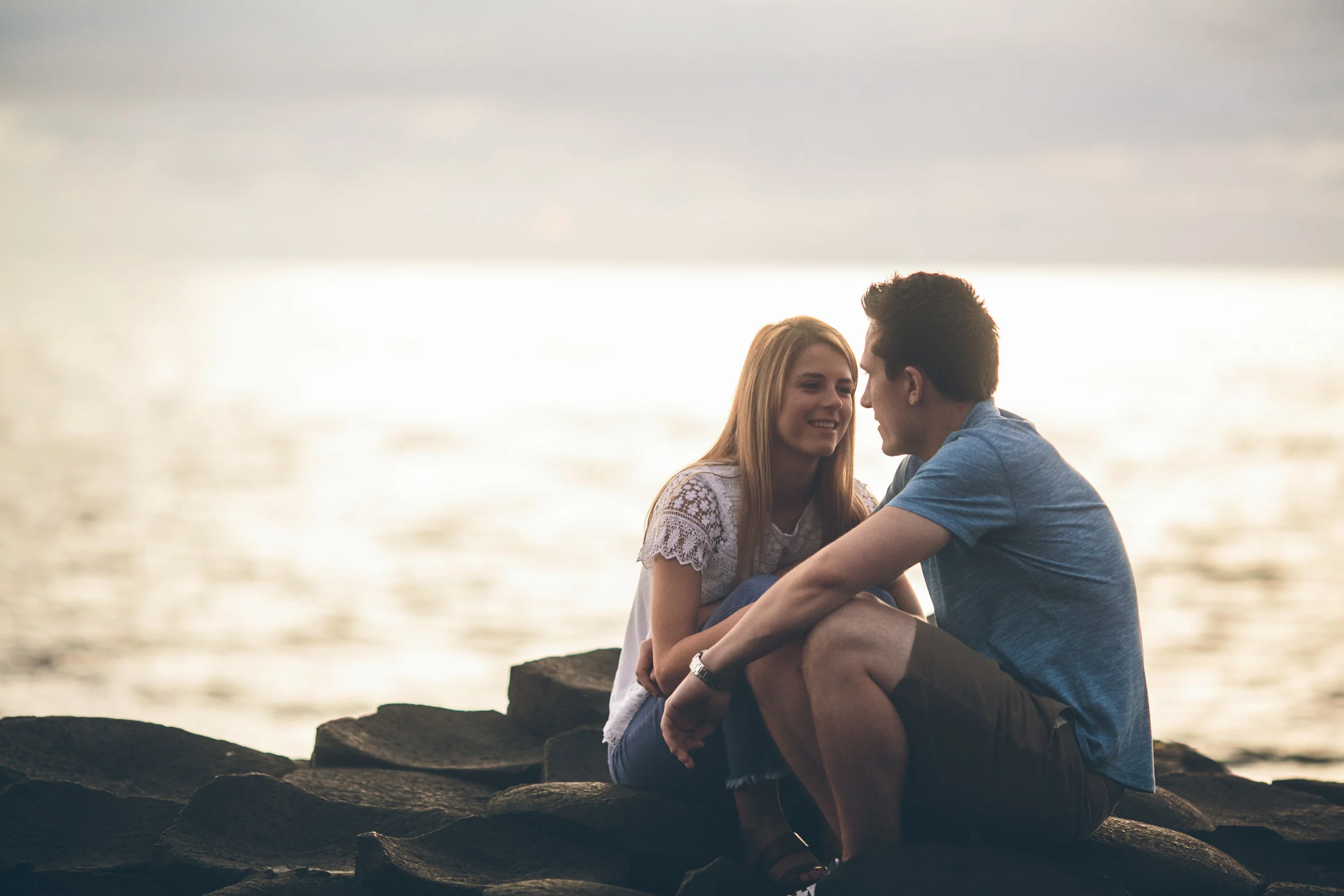 A young couple sitting on rocks by a body of water, facing each other and smiling at each other during sunset.