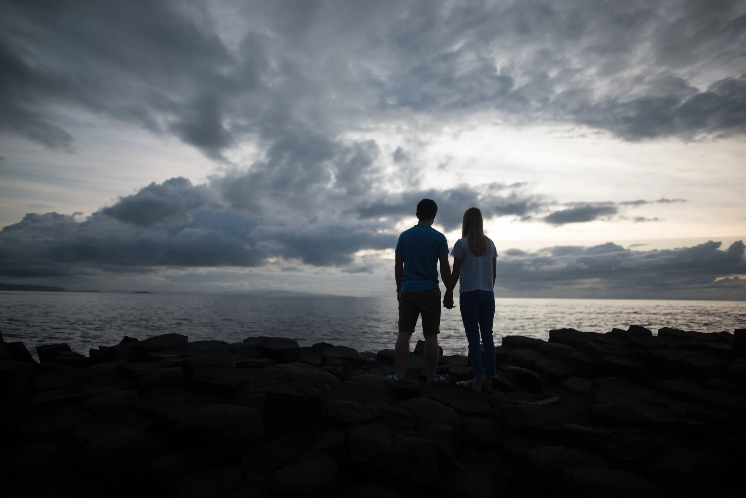 A couple holding hands and standing on rocks by the water, facing a cloudy sky at sunset or dusk.