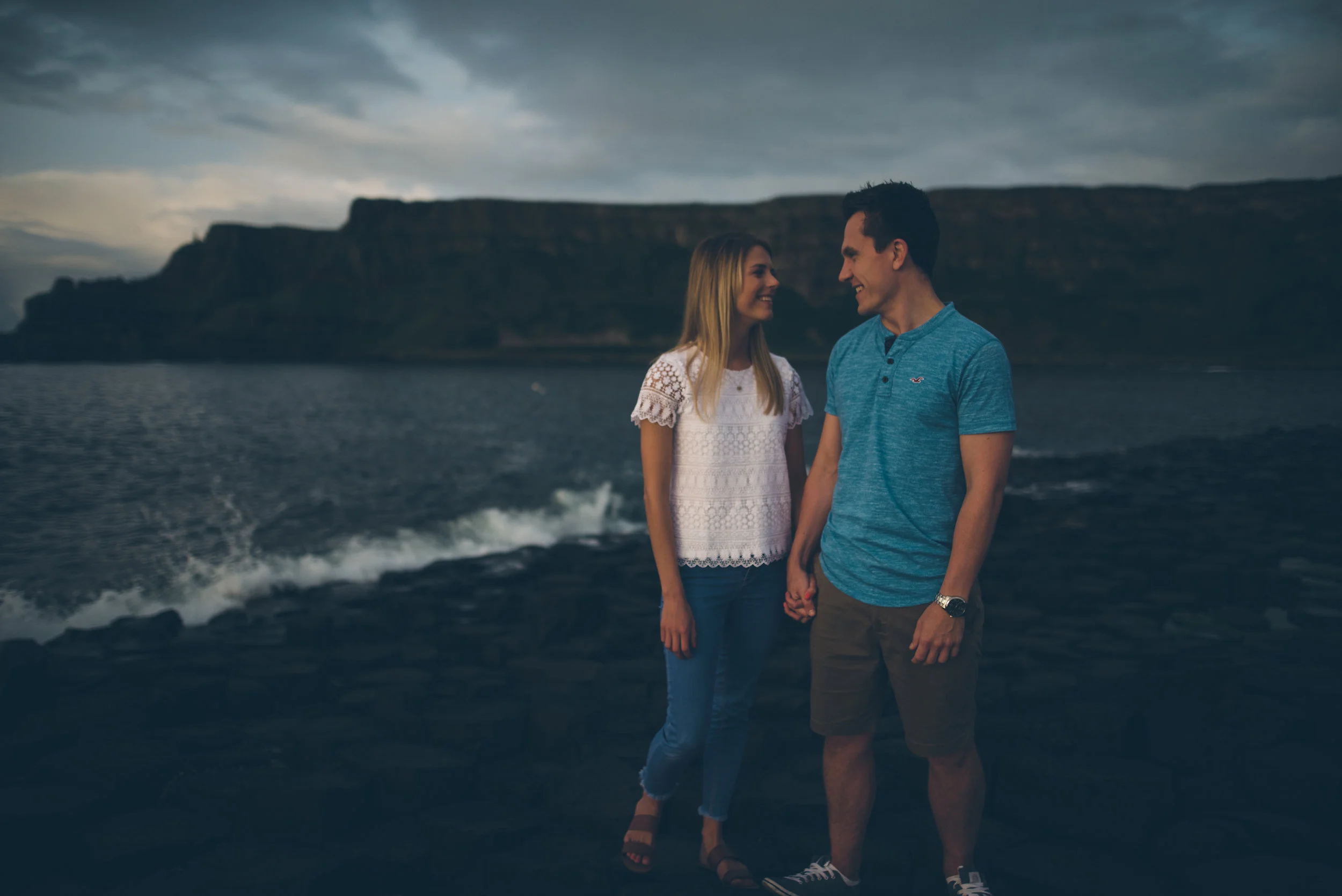 A couple holding hands and smiling at each other near a body of water with cliffs in the background during cloudy weather.