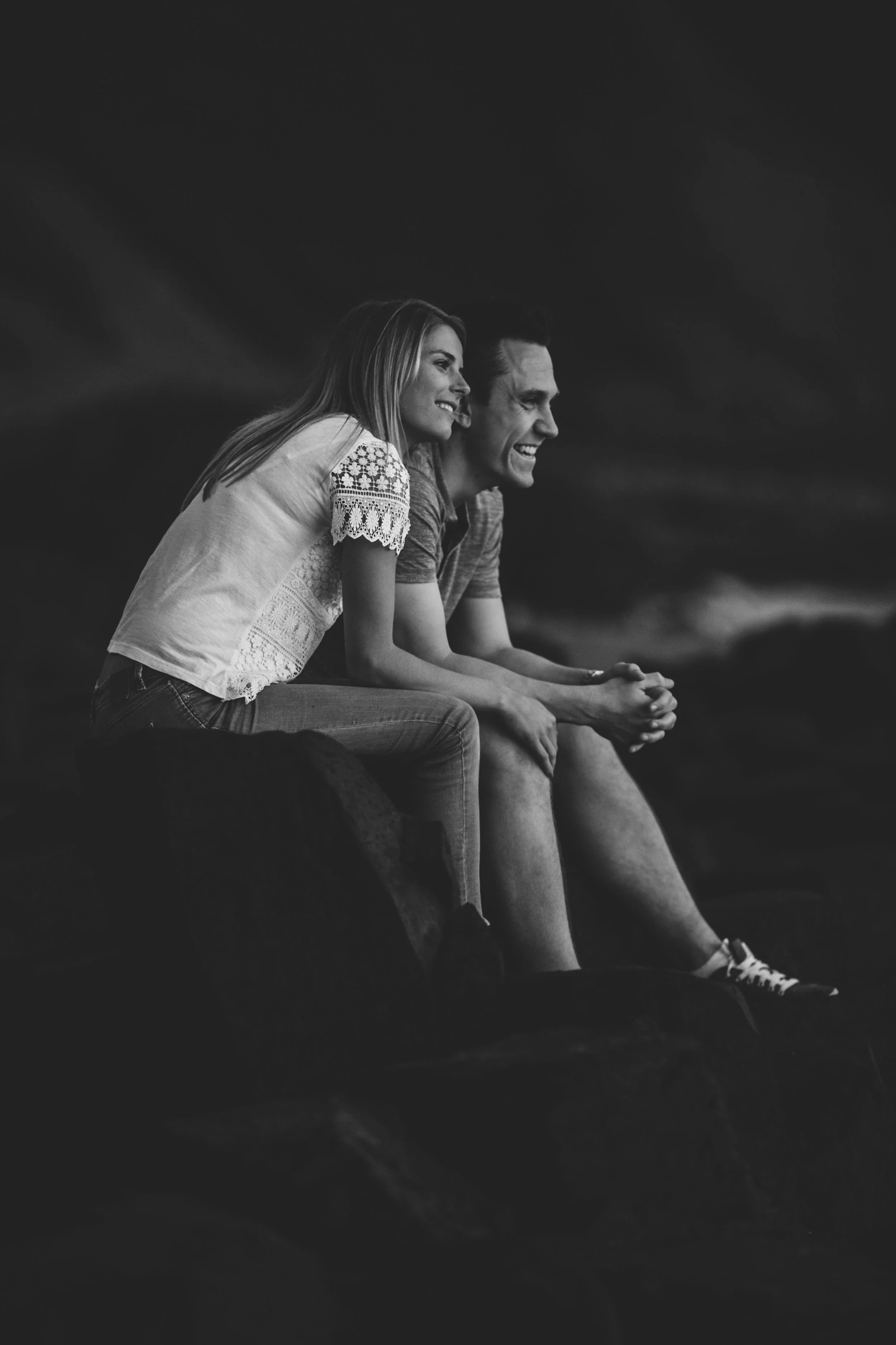 A black-and-white photo of a young woman and man sitting on rocks, smiling and looking to the right, with a dark background.