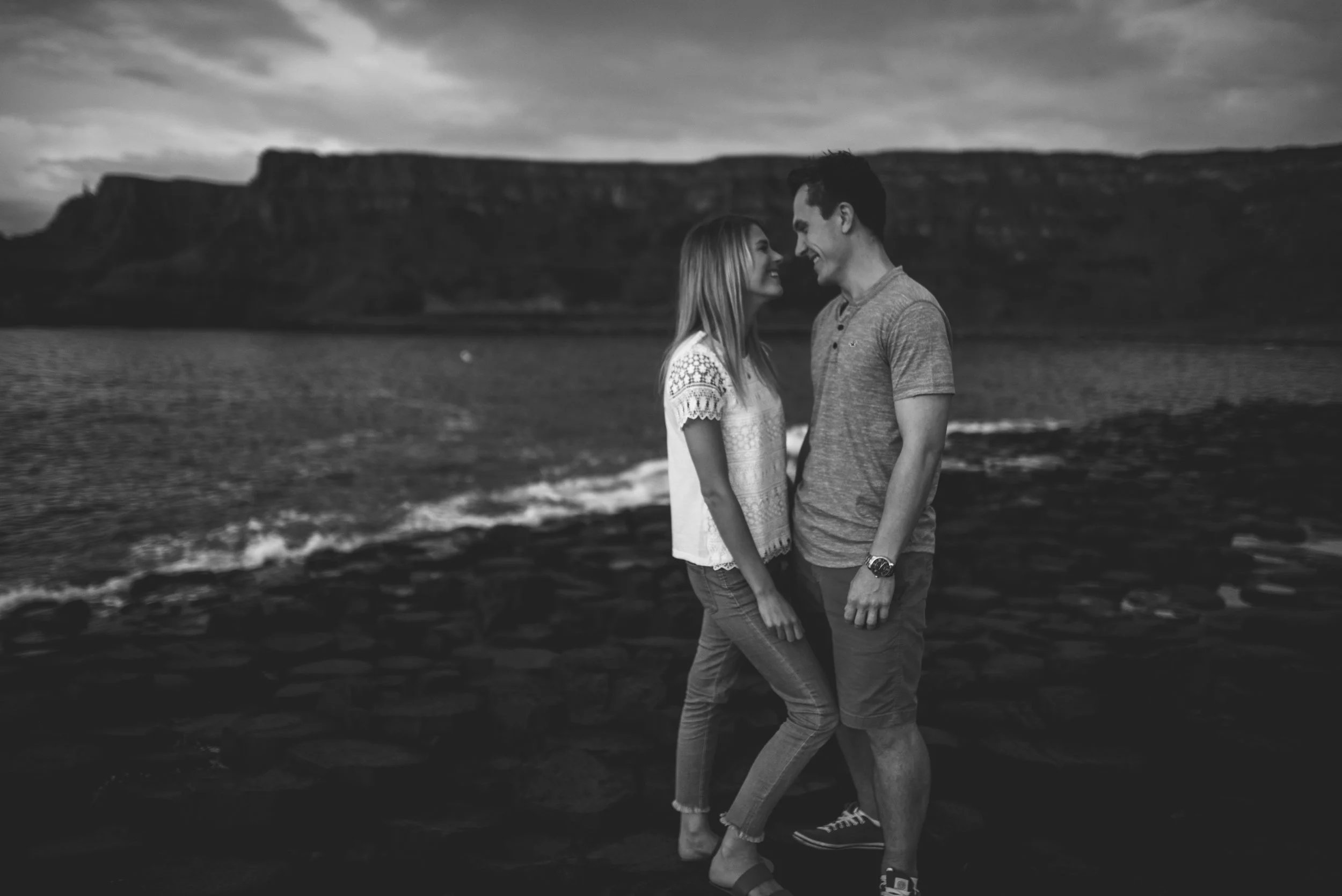 A black-and-white photo of a couple standing close together on a rocky beach, smiling and leaning towards each other with a mountain and cloudy sky in the background.