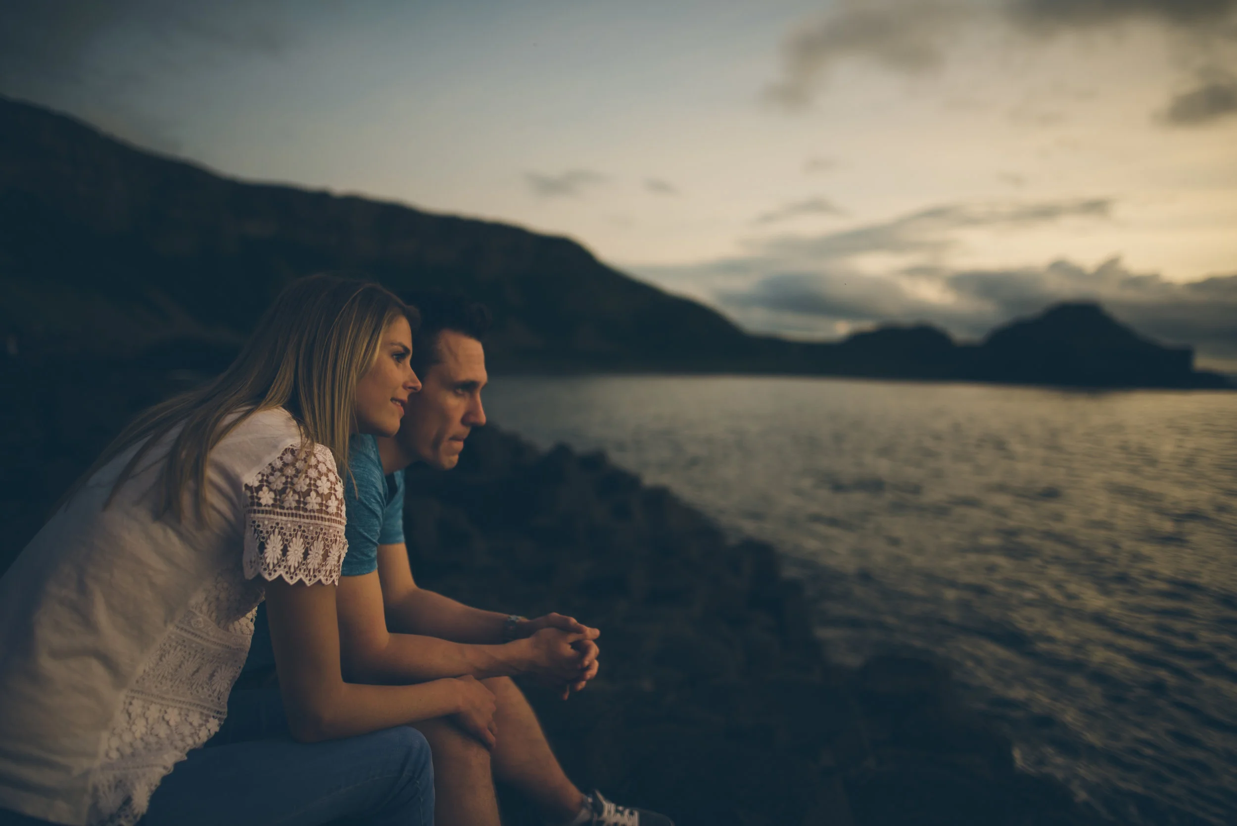 A man and woman sit on rocks by the water during sunset, looking contemplatively at the horizon with mountains in the background.