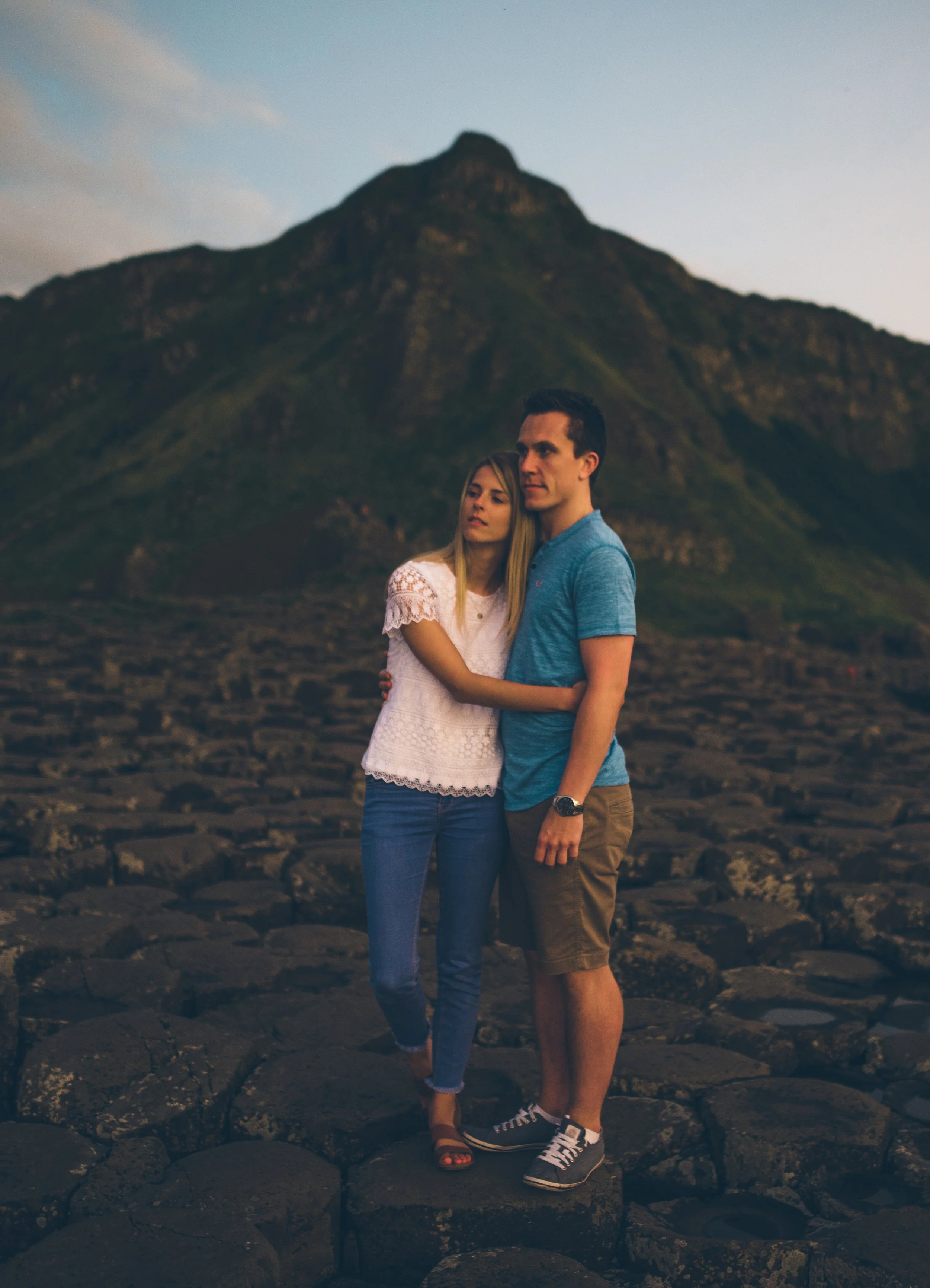 A couple standing on rocky terrain with a mountain in the background, during sunset or dusk. Giants Causeway

