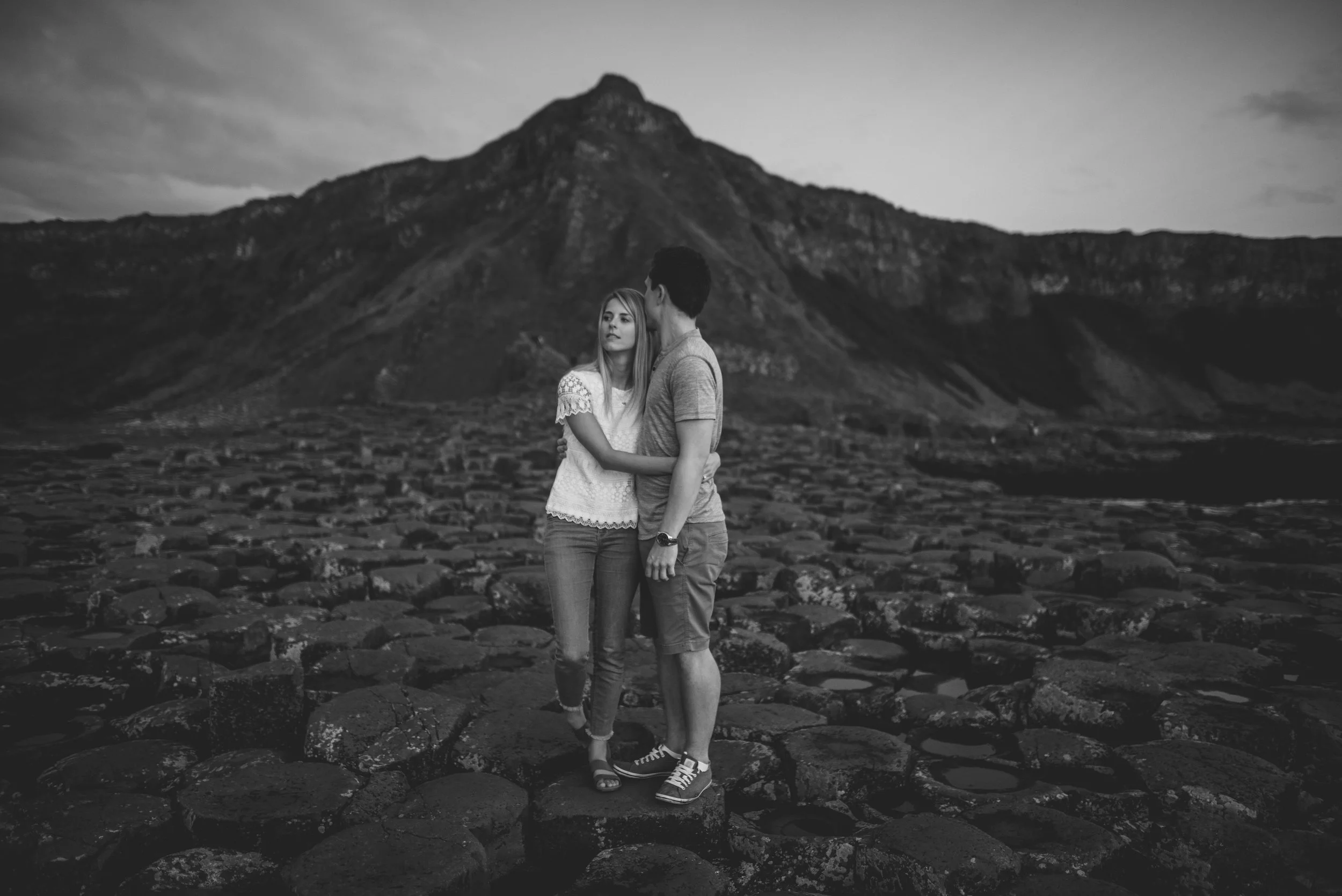 A couple standing together on a rocky landscape with a mountain in the background, in black and white. Giants Causeway