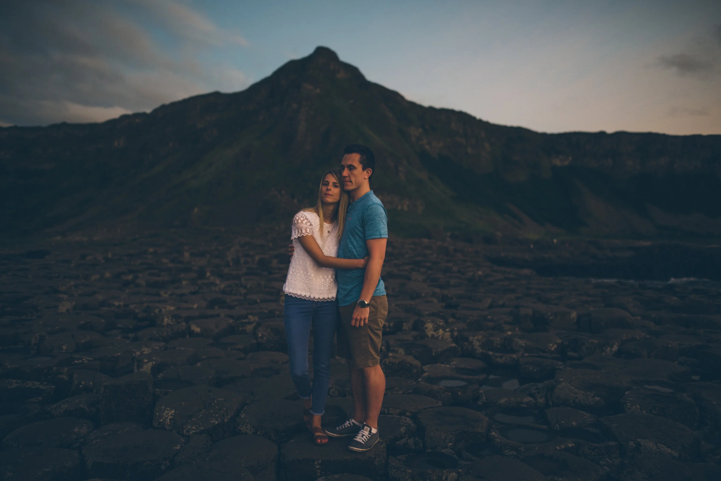 A young couple standing closely together on a rocky landscape with a mountain in the background, during what appears to be dusk or dawn. Giants Causeway