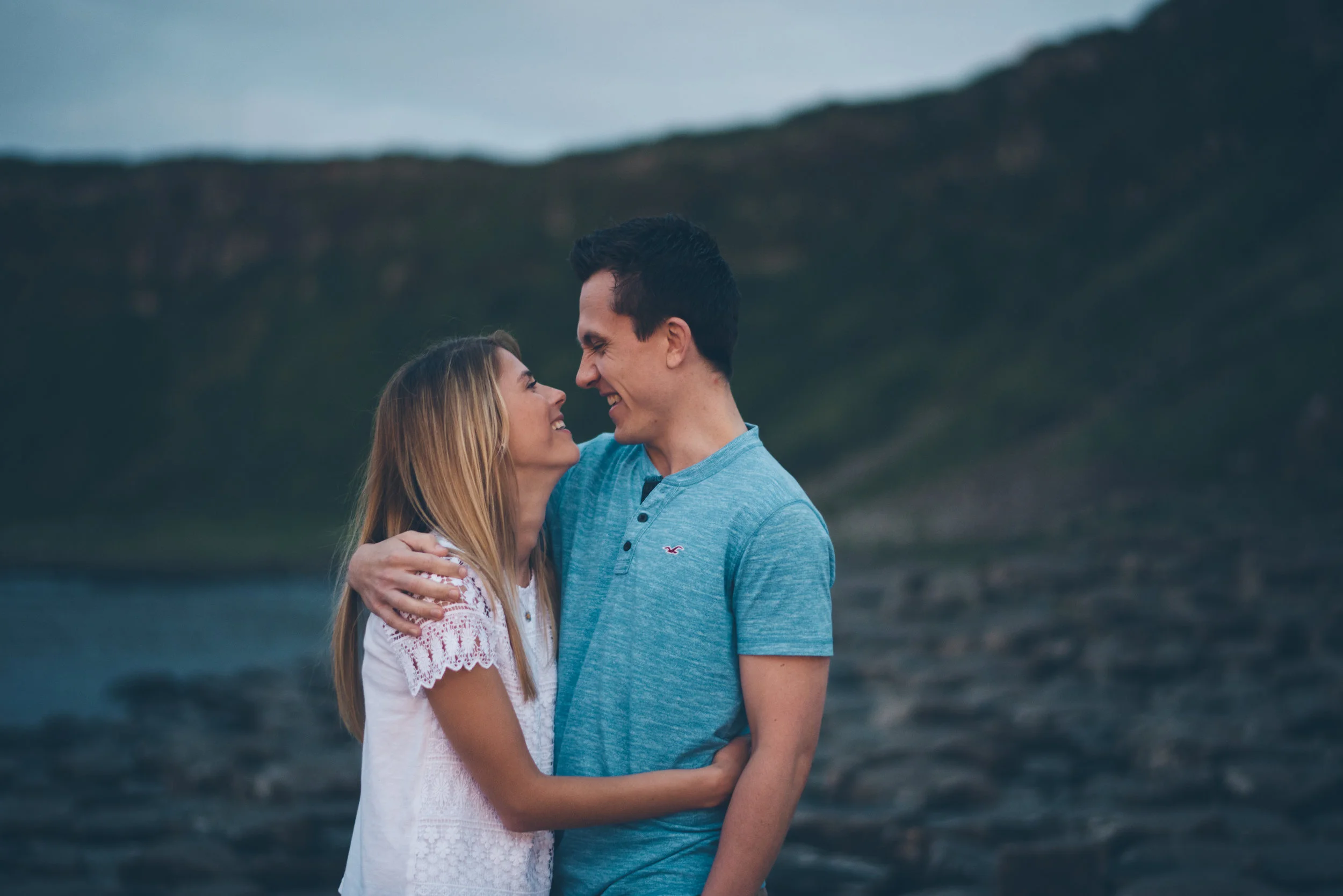 A young couple smiling and embracing outdoors near a body of water with hills in the background.