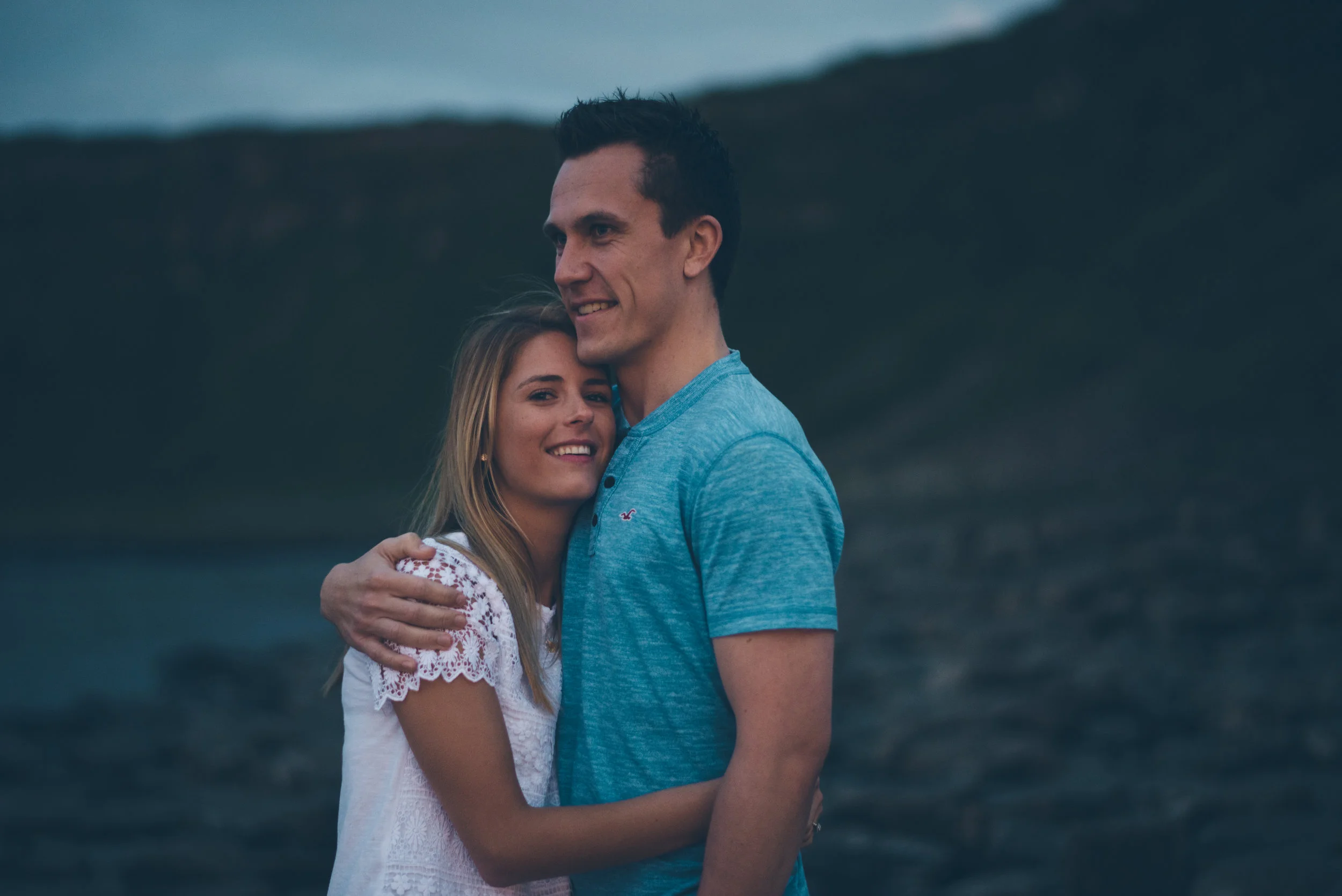 A smiling young woman and a tall man are hugging outdoors near a body of water with a mountain and cloudy sky in the background.