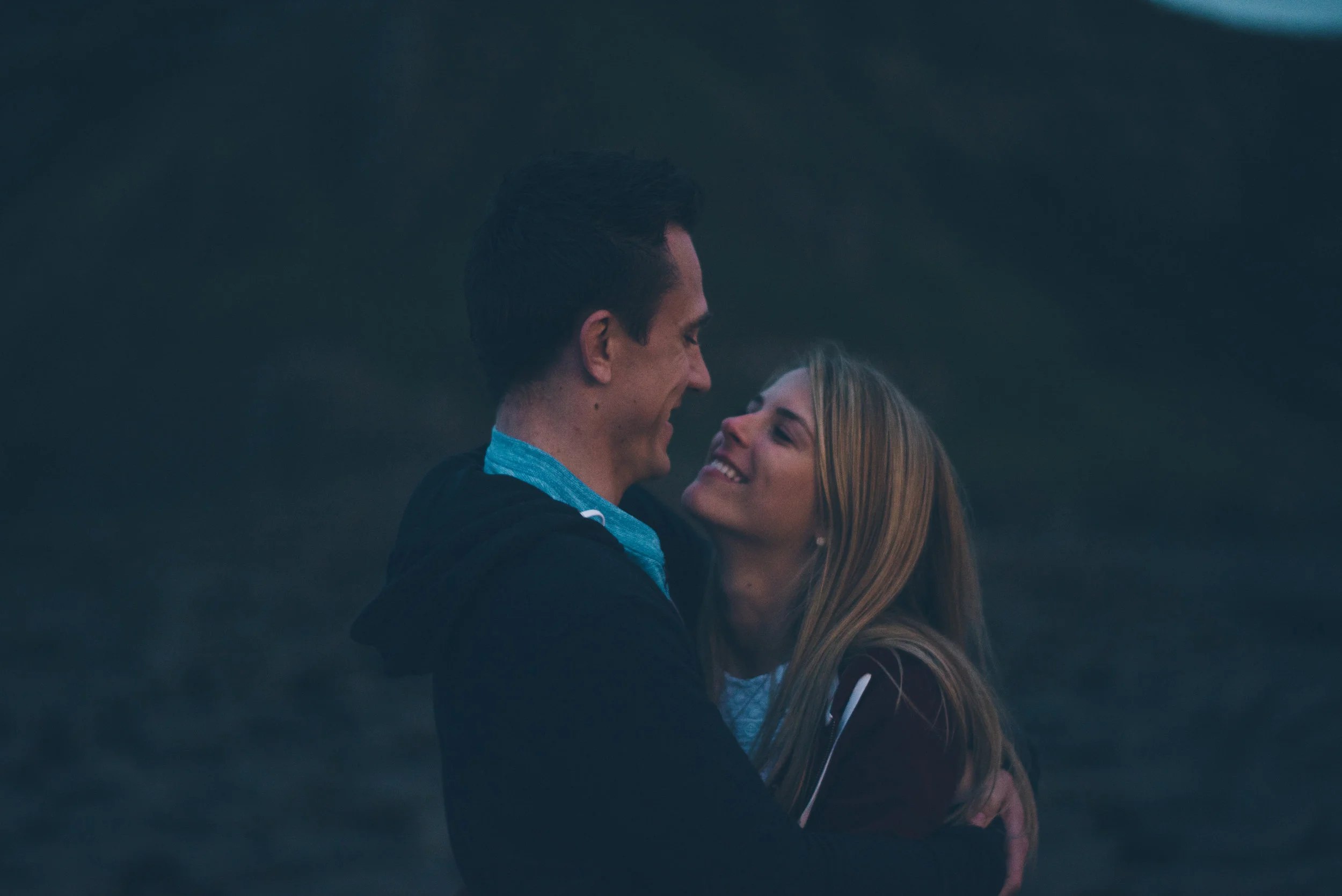 A young man and woman embrace outdoors at dusk, looking at each other and smiling, with blurred dark background.