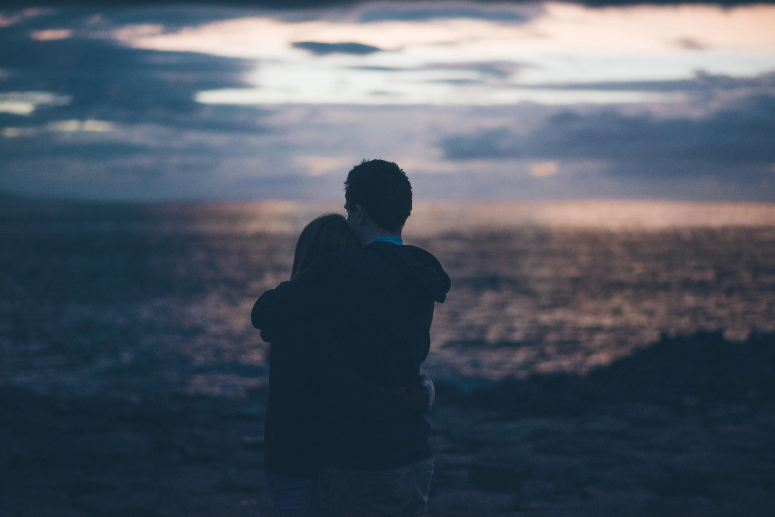 A couple hugging at the beach during sunset or twilight, with the ocean and cloudy sky in the background.