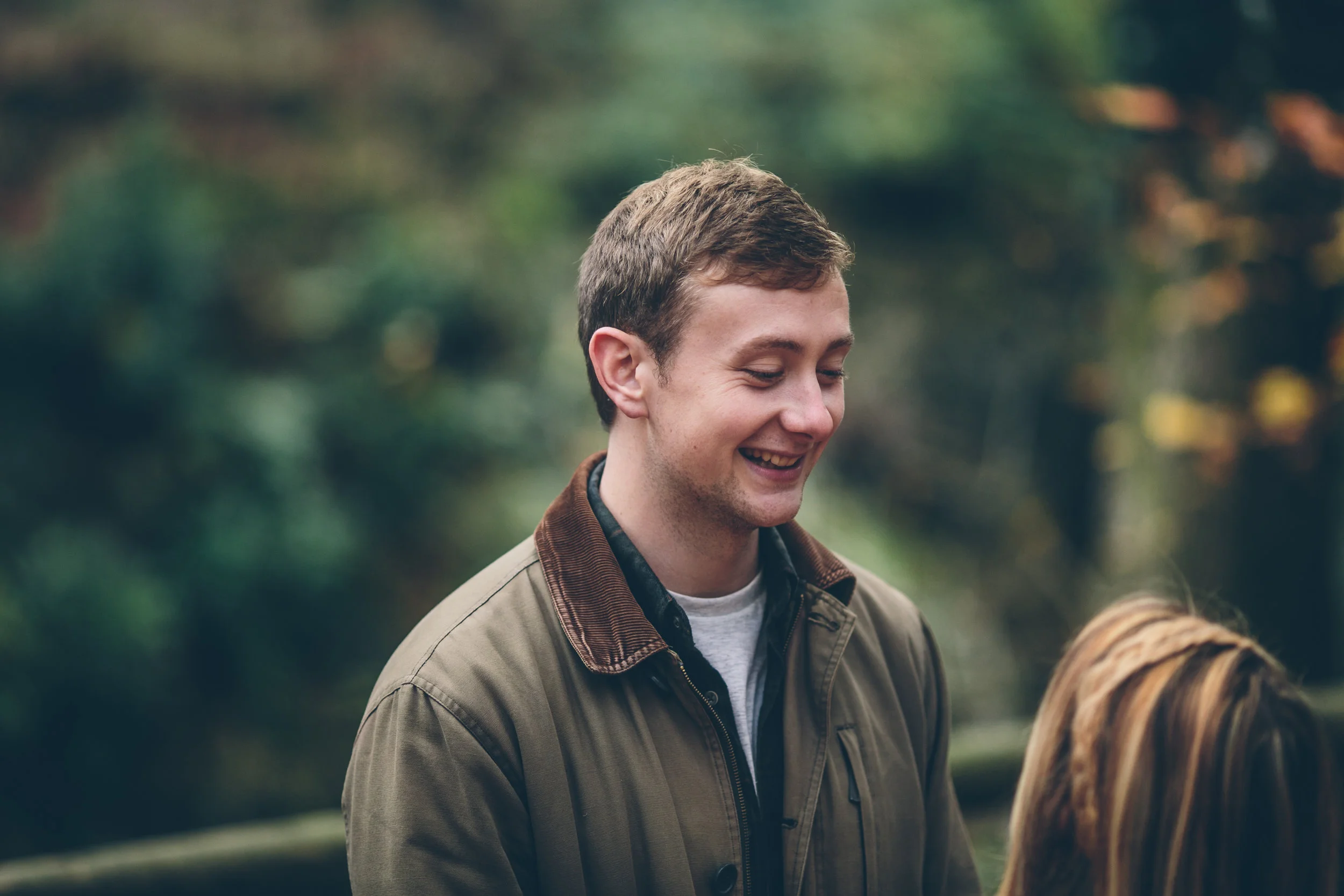 A young man with short brown hair and a light complexion is smiling while engaged in conversation with someone whose head with blonde hair is partially visible in the foreground. They are outdoors with blurred greenery and trees in the background.