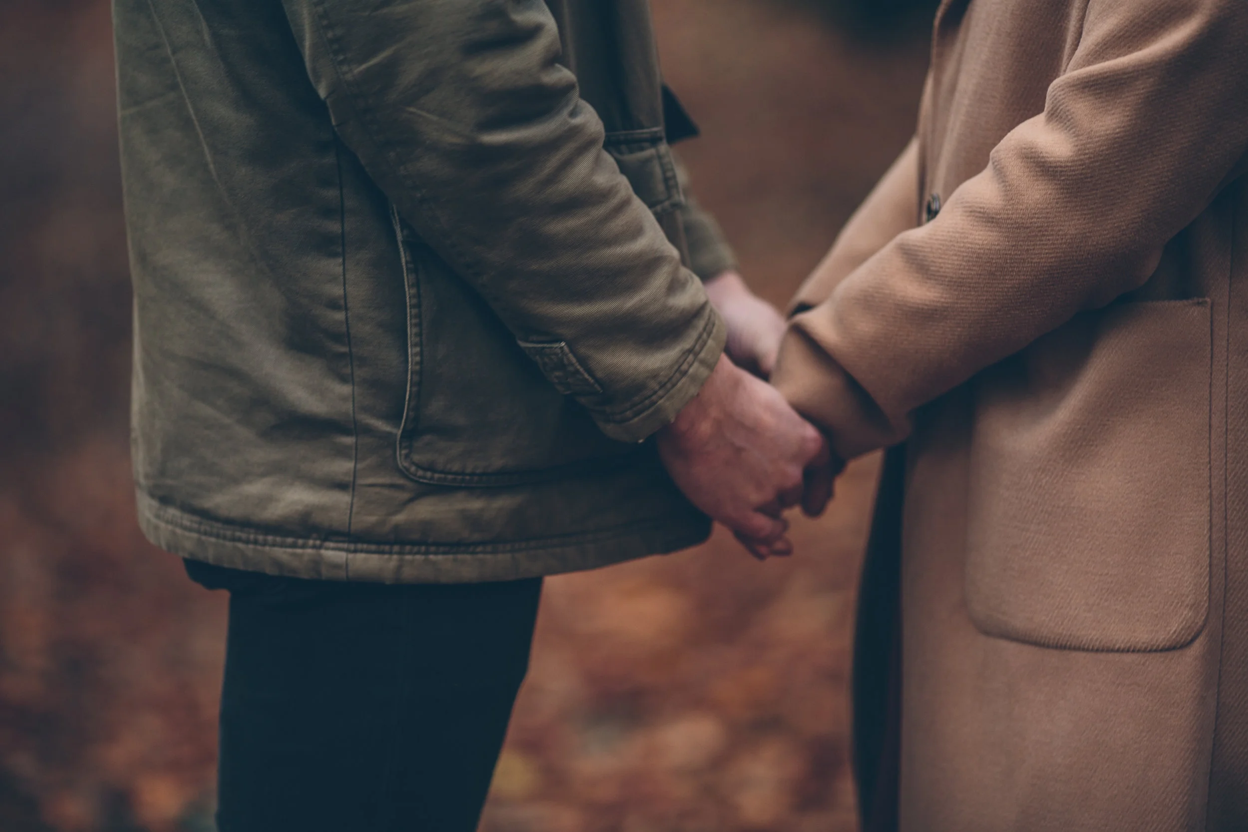 Close-up of two people holding hands, wearing jackets, outdoors with a blurred background of fallen leaves.