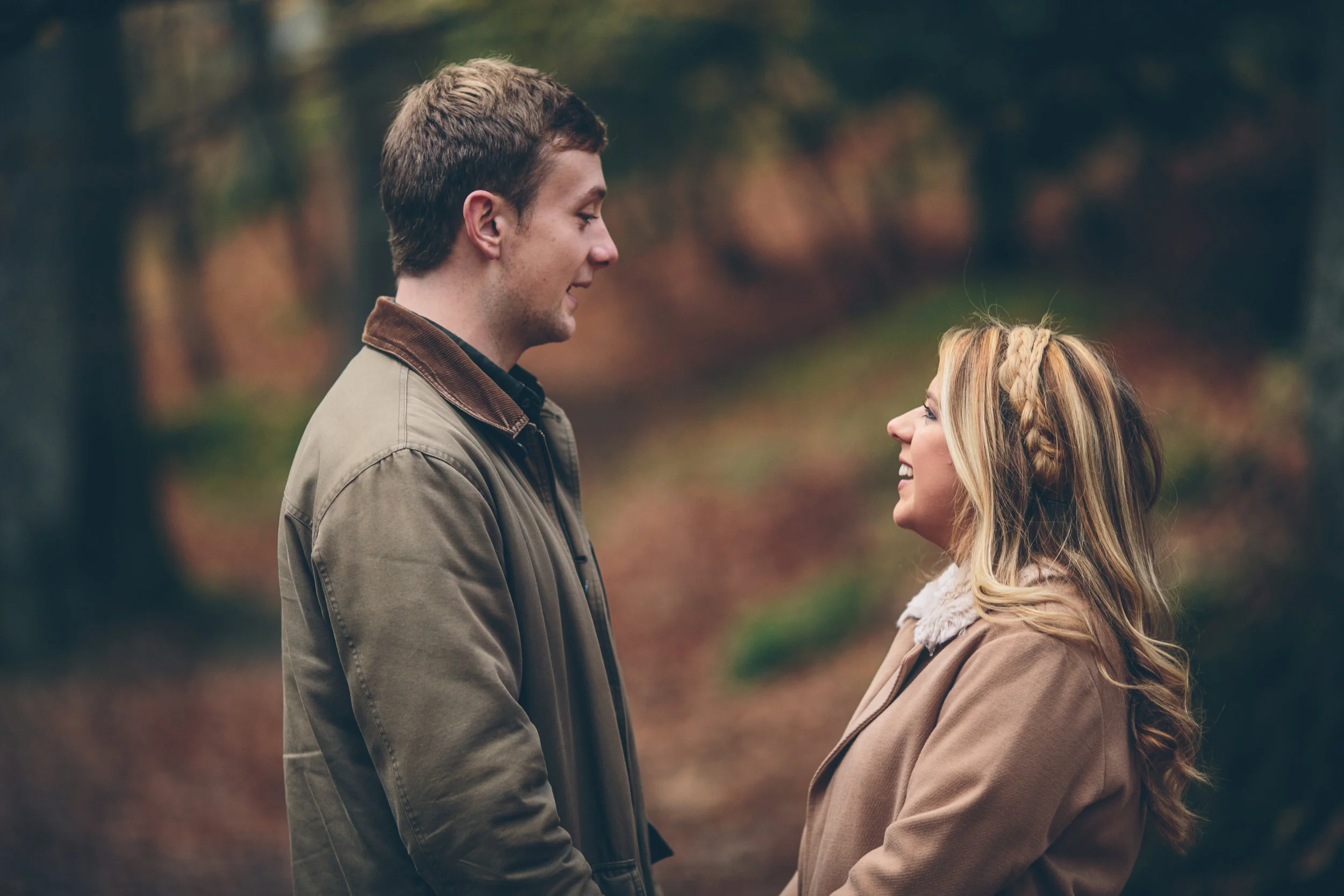 A young man and woman stand facing each other outdoors in a forest or park during autumn, smiling and looking into each other's eyes.