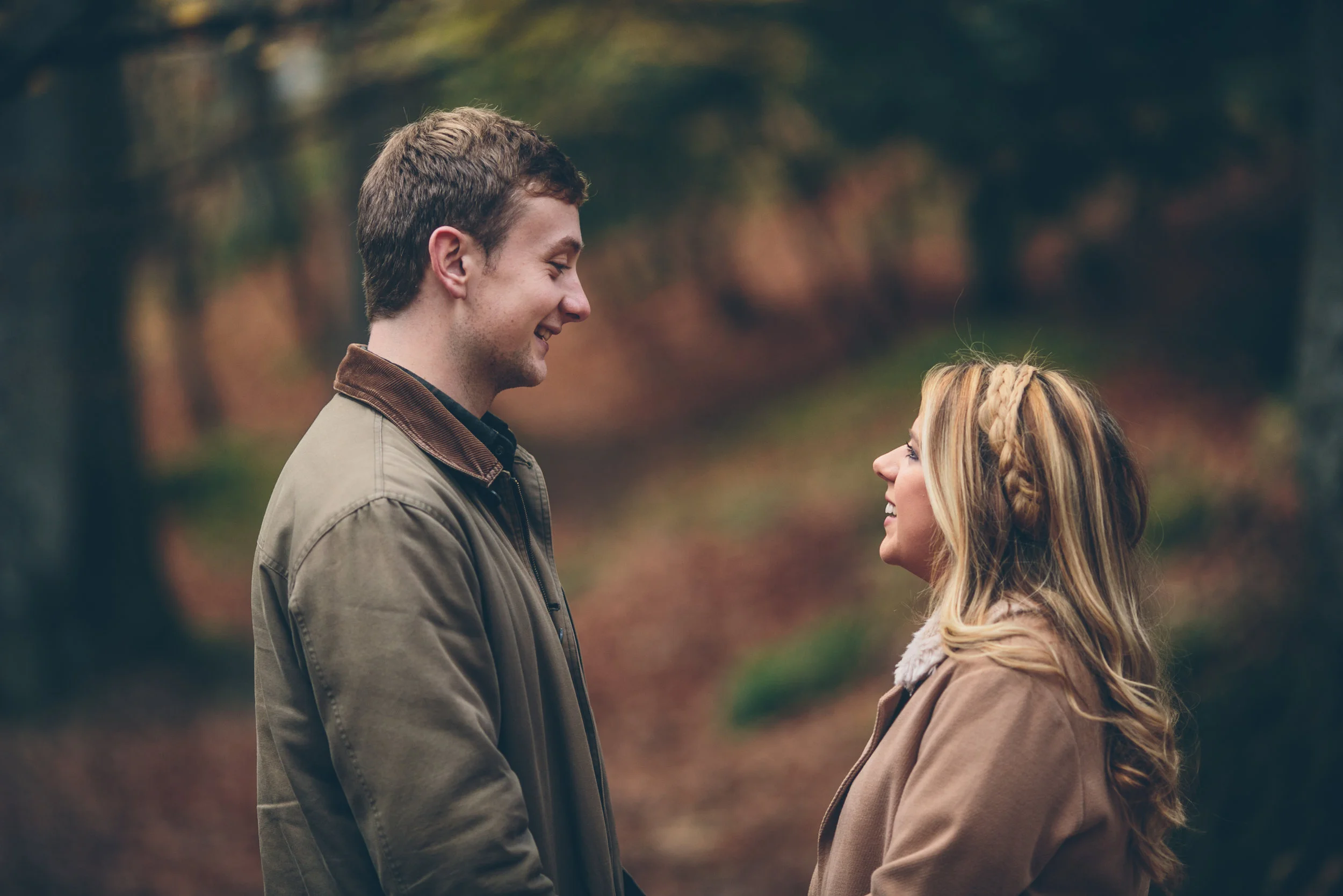 A young man and woman standing outdoors in a forest, facing each other and smiling.
