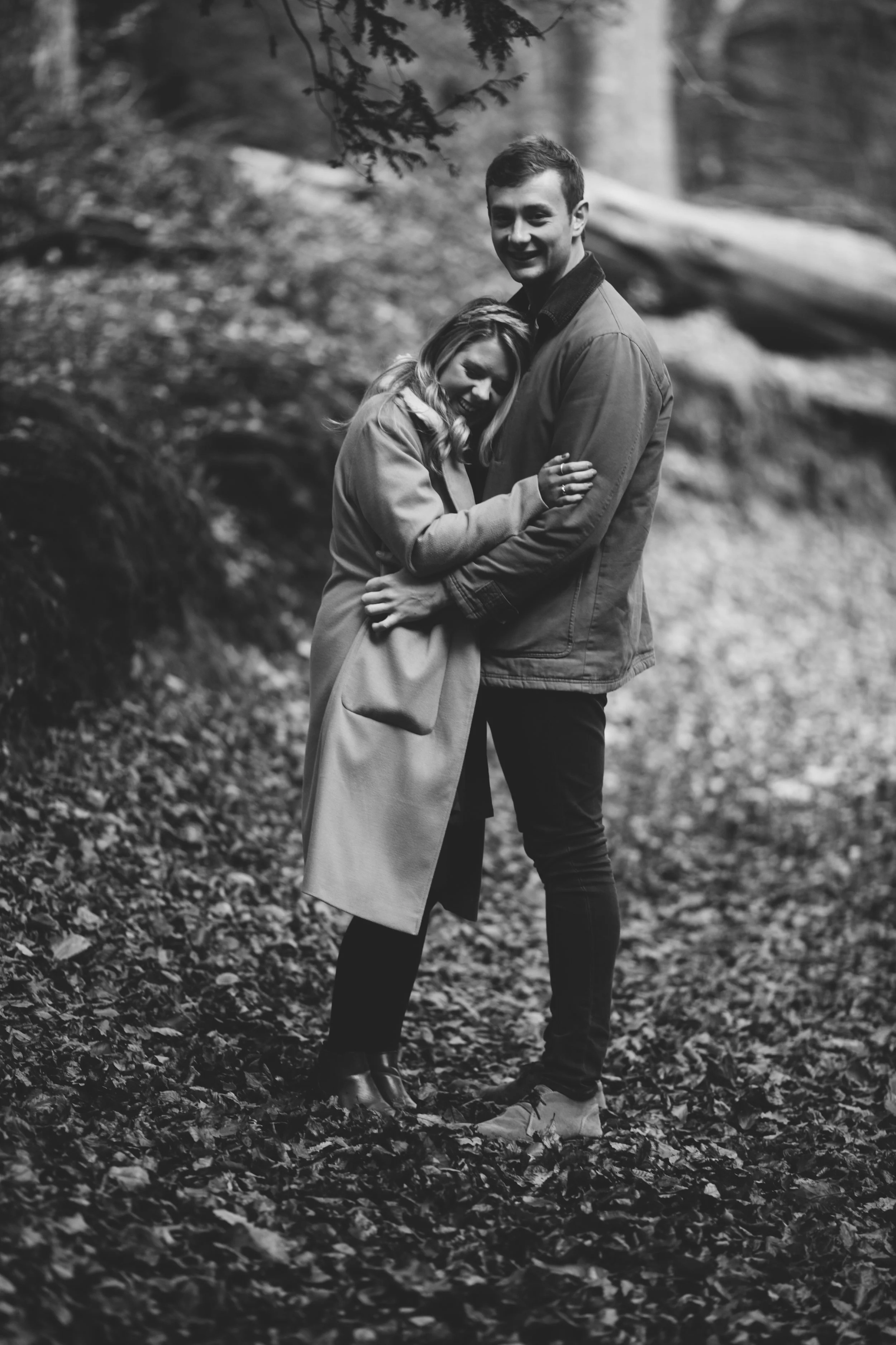 Black and white photo of a couple hugging outdoors on a leaf-covered ground, with trees in the background.