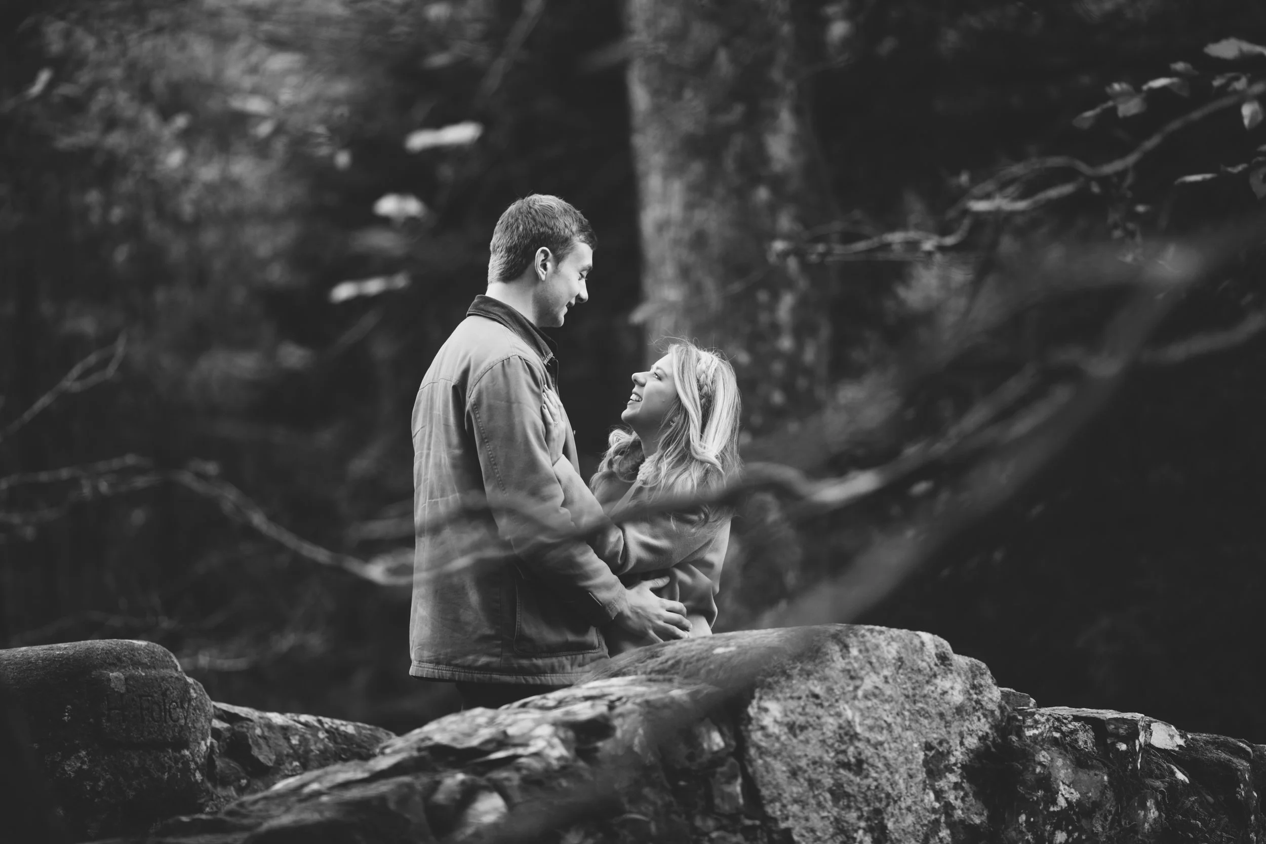 A black and white photo of a couple standing on a rock outdoors, smiling and looking at each other, surrounded by trees and forest.
