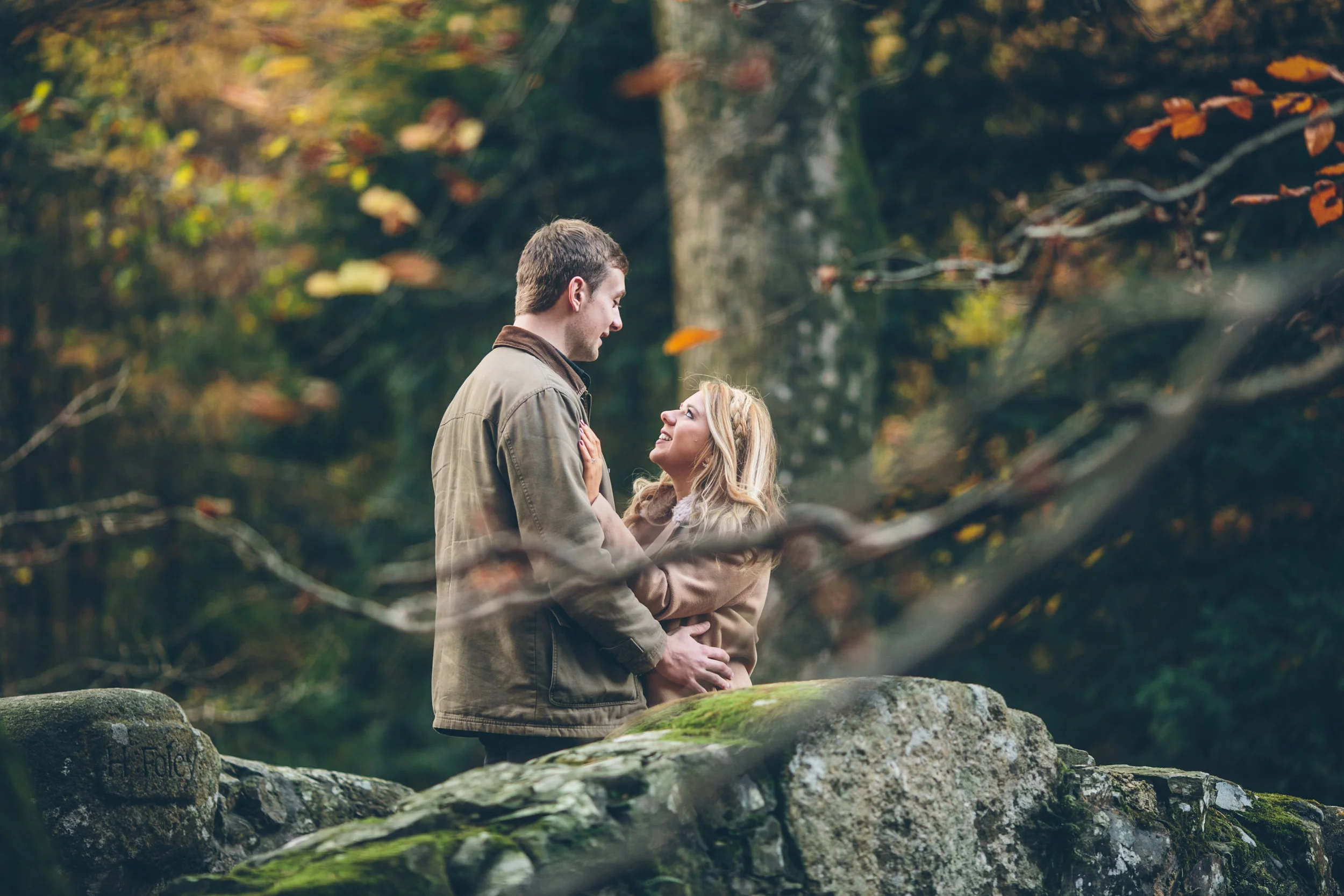 A man and a woman looking at each other affectionately, standing on a moss-covered rock in a wooded area with autumn leaves. The woman has blonde hair and wears a beige coat, while the man has brown hair and wears a brown jacket.