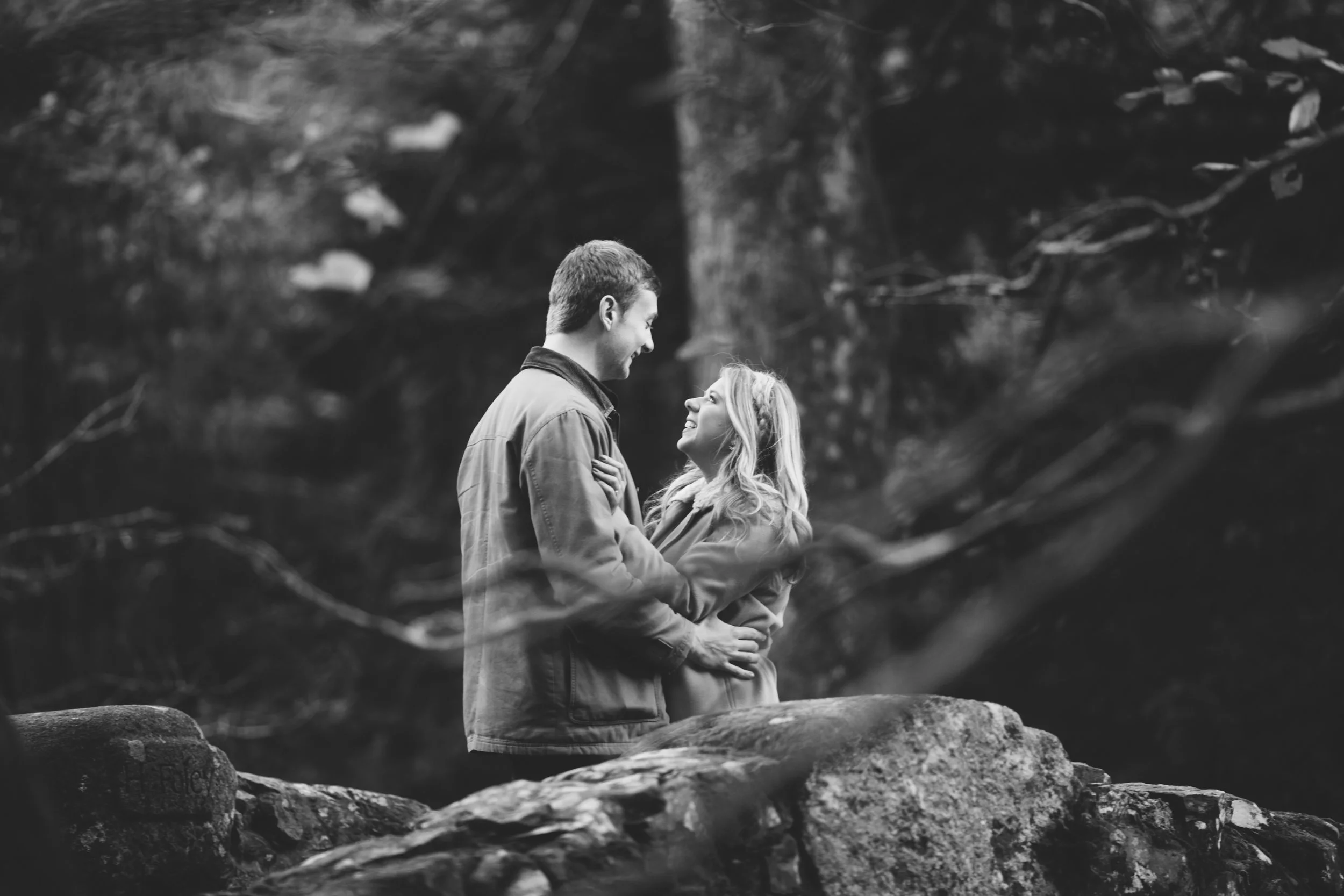 A black-and-white photo of a couple standing close together on a large rock, looking into each other's eyes and smiling, surrounded by a natural outdoor setting.