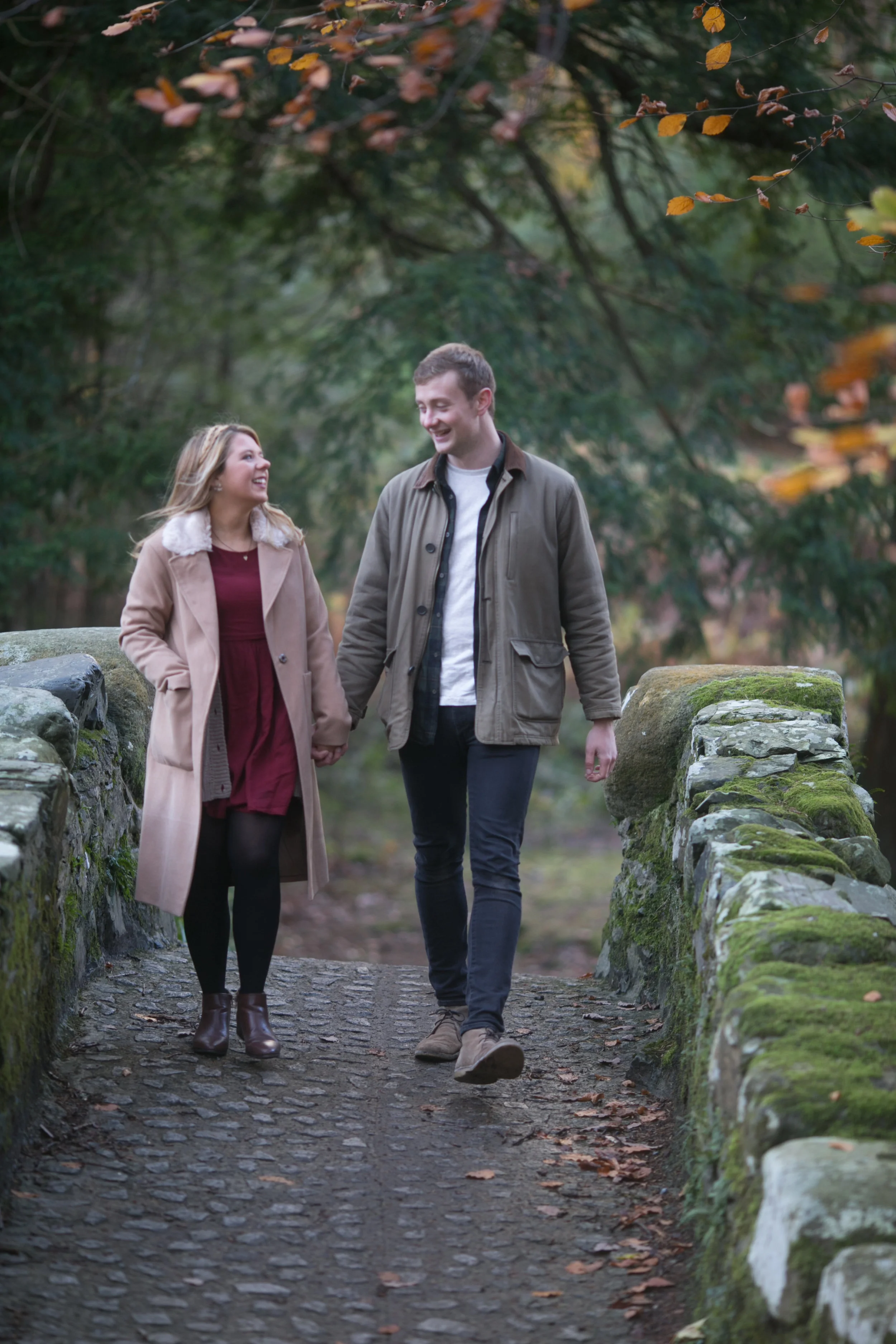 A young couple walking hand-in-hand on a stone pathway with moss-covered stone railing, surrounded by autumn trees, smiling and enjoying a fall day.