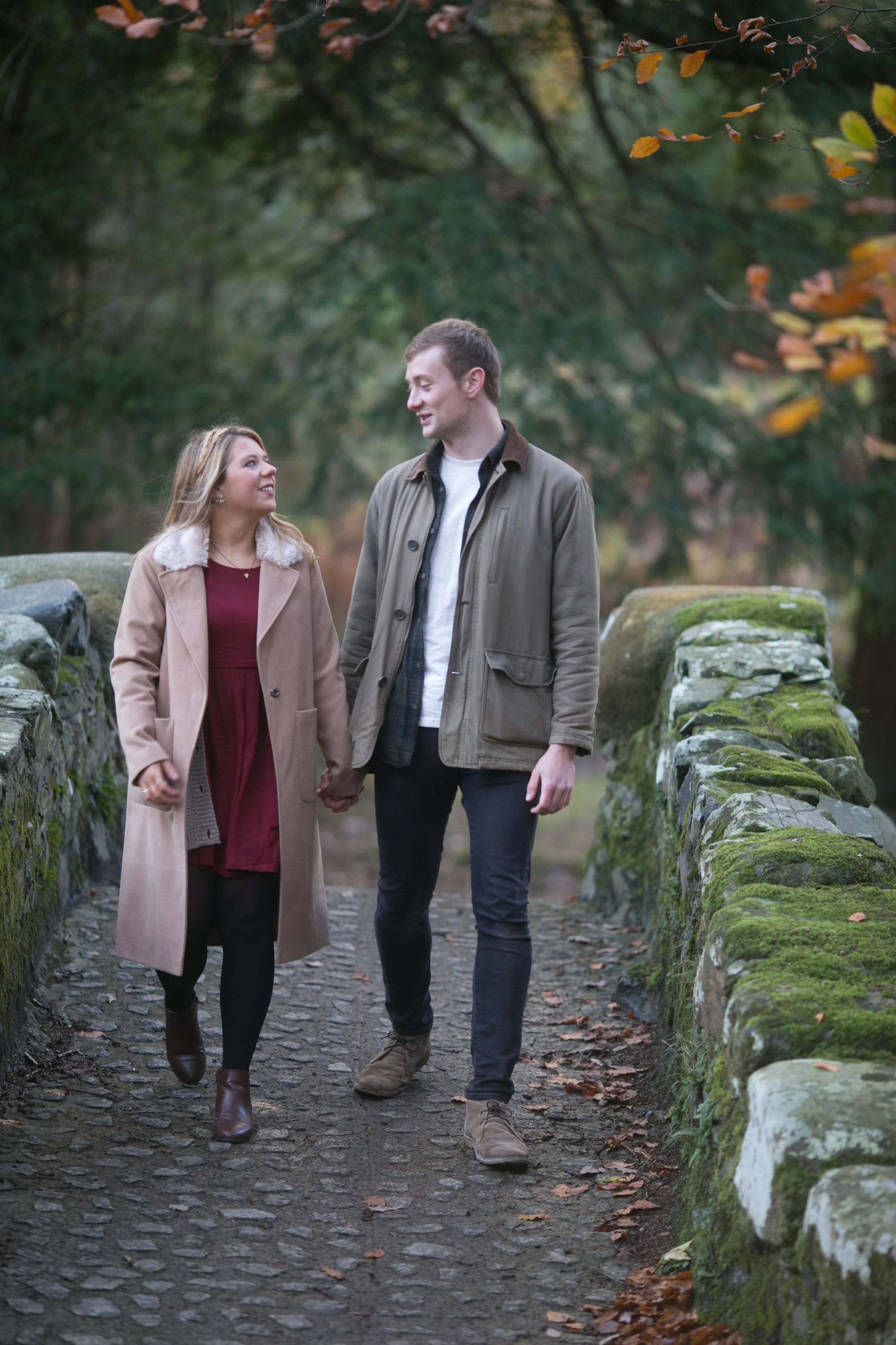 A young couple holding hands and walking on a stone path surrounded by moss-covered rocks and trees.