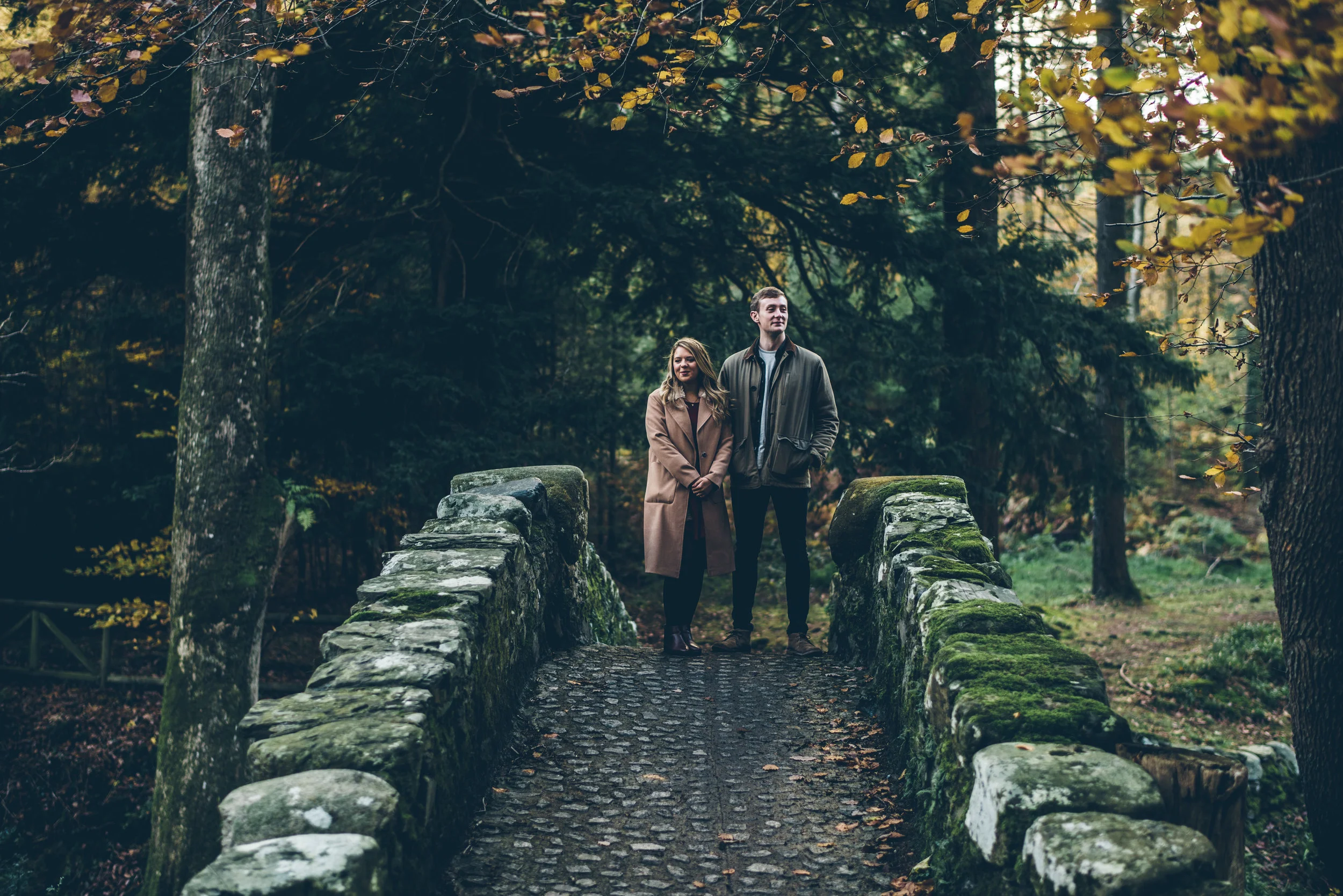 A man and a woman standing on a small stone bridge in a forest with autumn leaves.