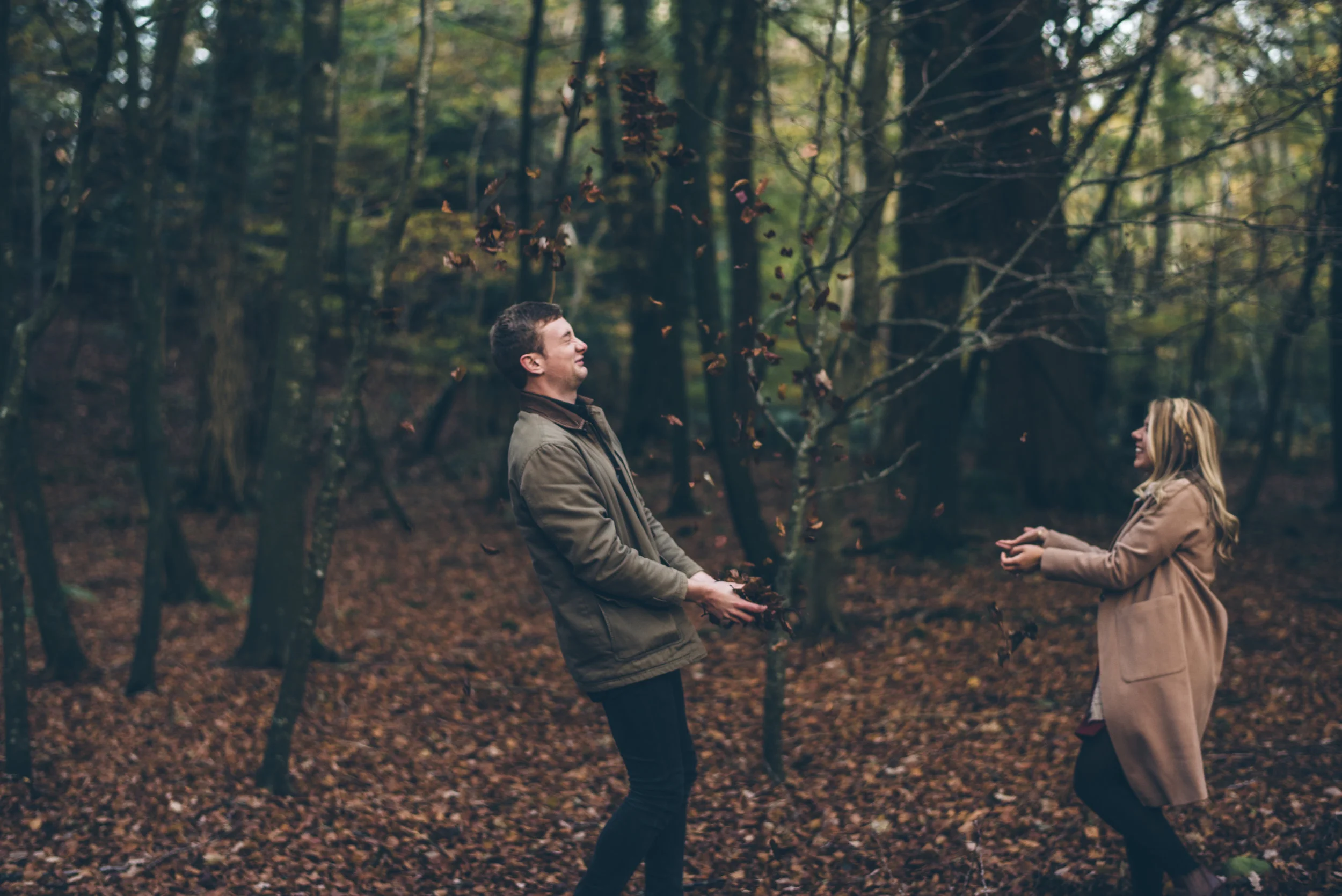 A man and woman playing with falling leaves in a wooded area during autumn.