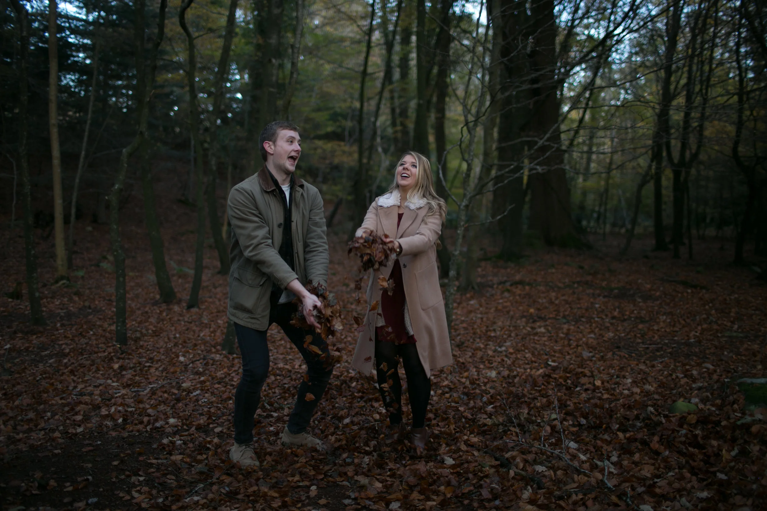A man and woman laughing and playing with fall leaves in a wooded forest, both holding leaves and dressed in jackets and coat.